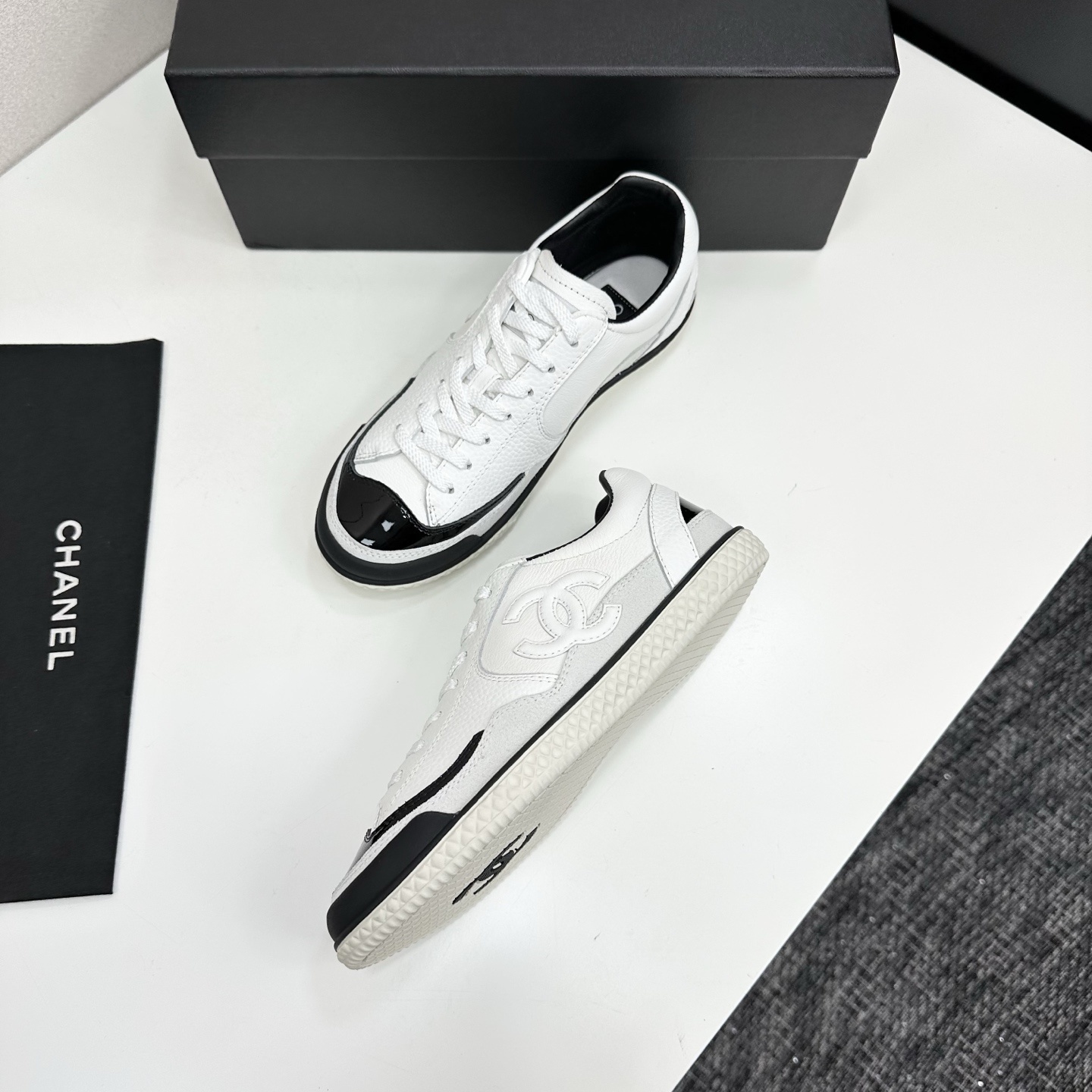 Side view of white Chanel calfskin sneakers with black patent leather details and white interlocking CC logo on a black box. - Hình 6