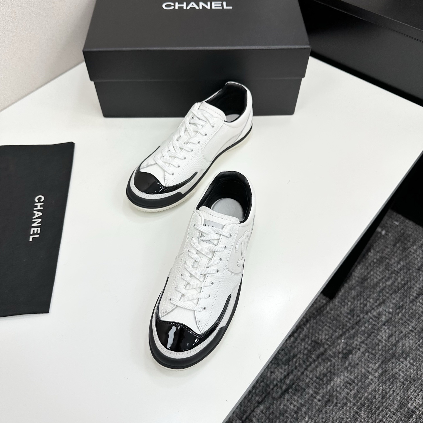 Side view of white Chanel calfskin sneakers with black patent leather details and white interlocking CC logo on a black box. - Hình 5