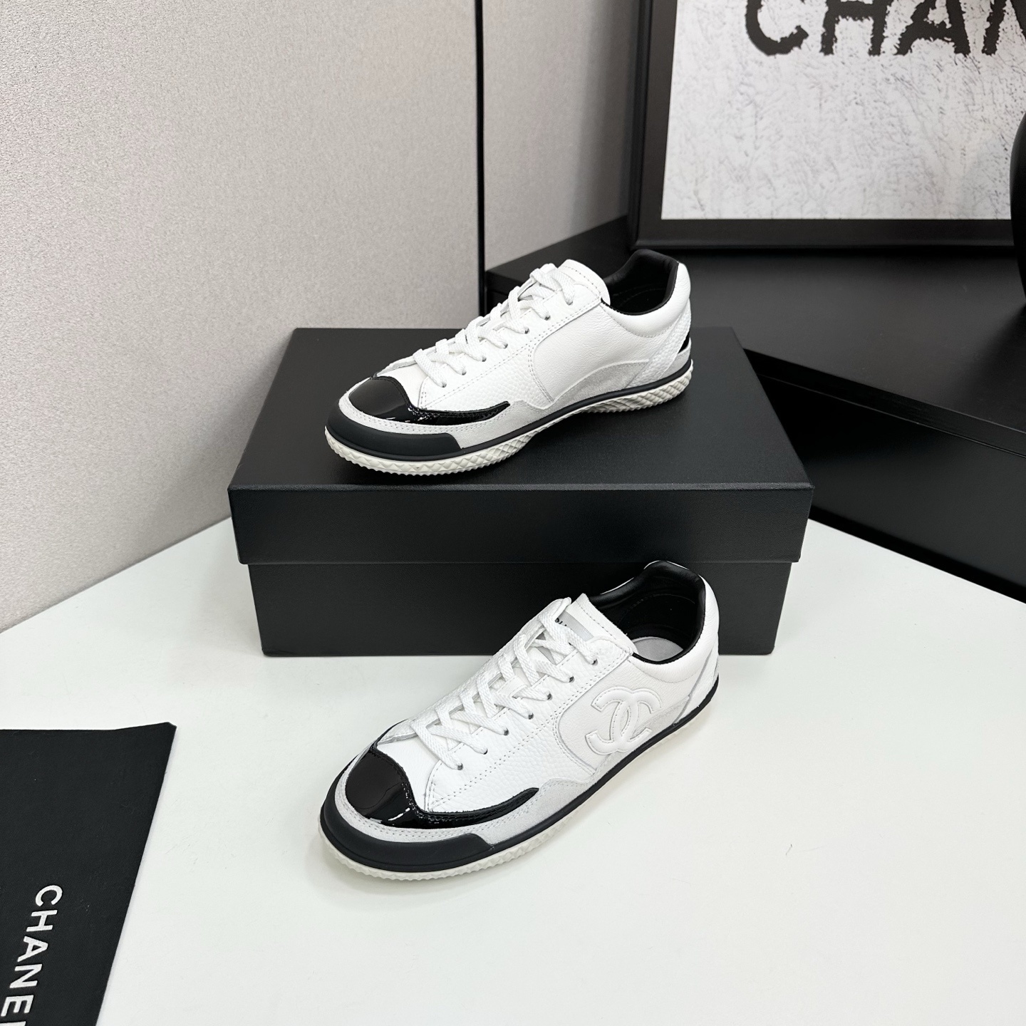 Side view of white Chanel calfskin sneakers with black patent leather details and white interlocking CC logo on a black box. - Hình 2