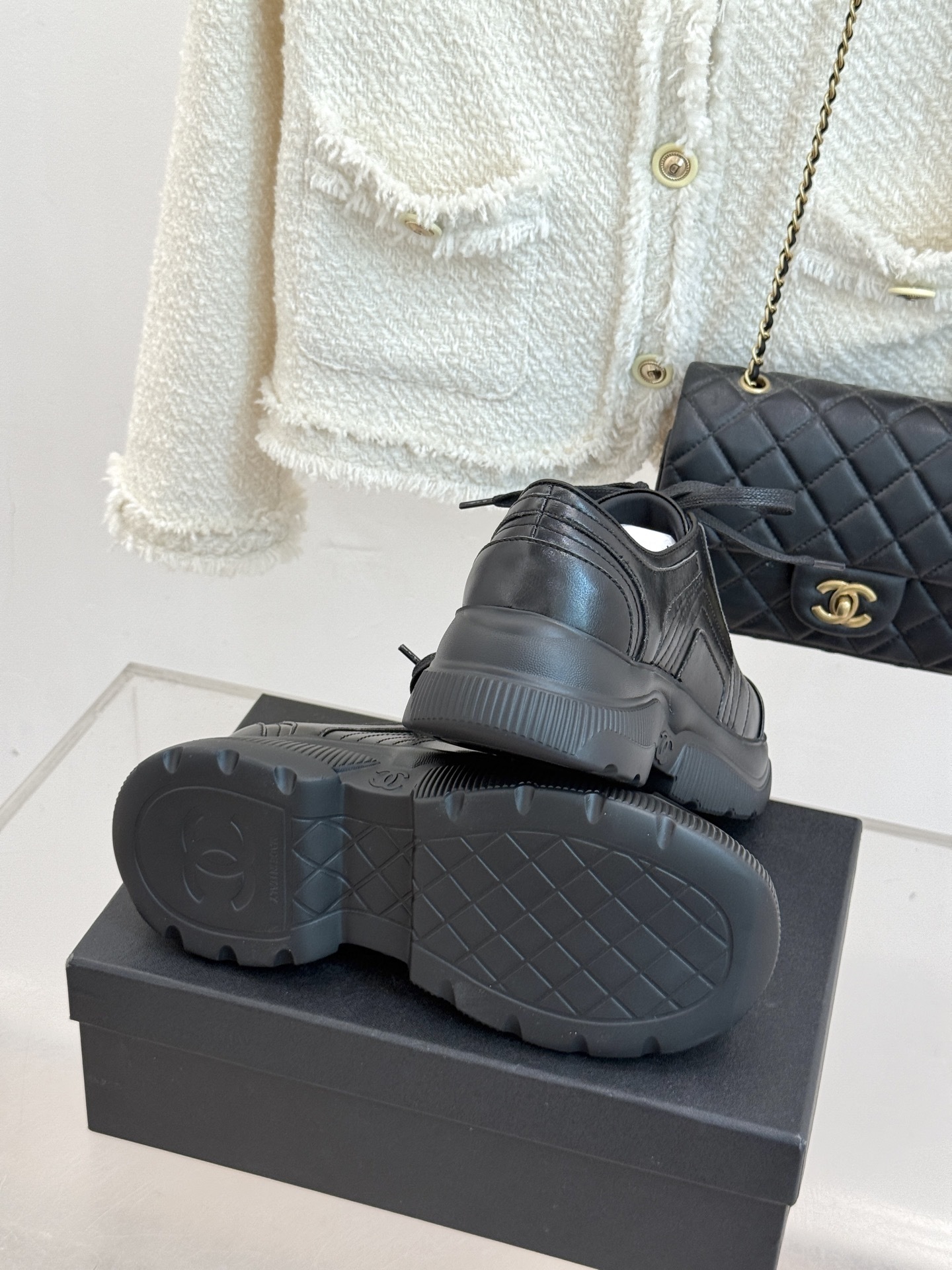A pair of black Chanel leather platform sneakers with embossed CC logo on a black box, styled with a white tweed jacket and black quilted bag. - Hình 9