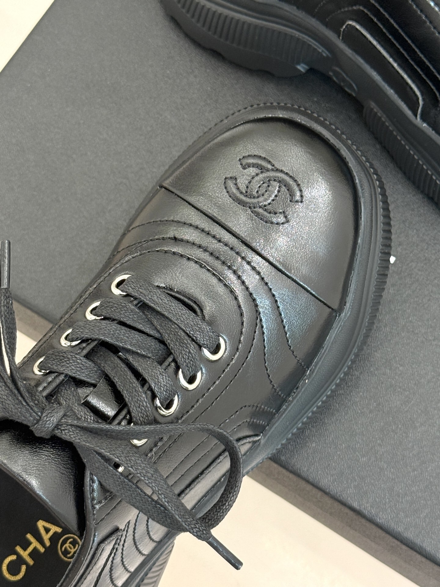 A pair of black Chanel leather platform sneakers with embossed CC logo on a black box, styled with a white tweed jacket and black quilted bag. - Hình 7