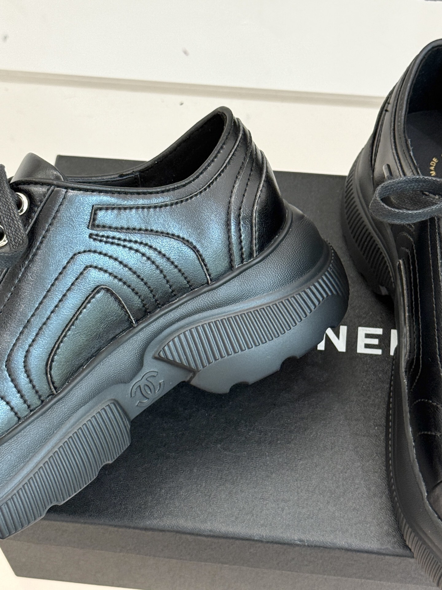 A pair of black Chanel leather platform sneakers with embossed CC logo on a black box, styled with a white tweed jacket and black quilted bag. - Hình 6