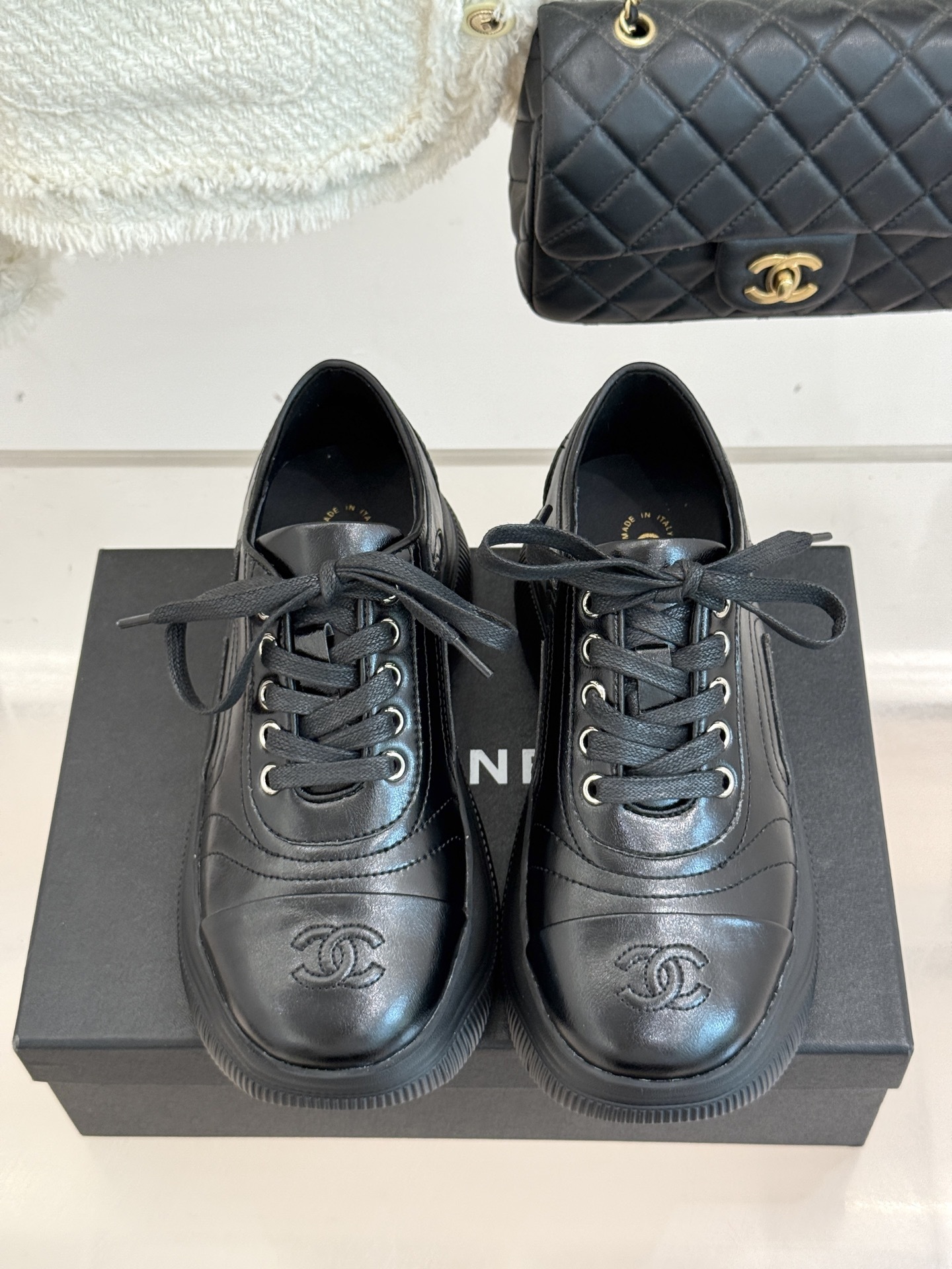 A pair of black Chanel leather platform sneakers with embossed CC logo on a black box, styled with a white tweed jacket and black quilted bag. - Hình 4