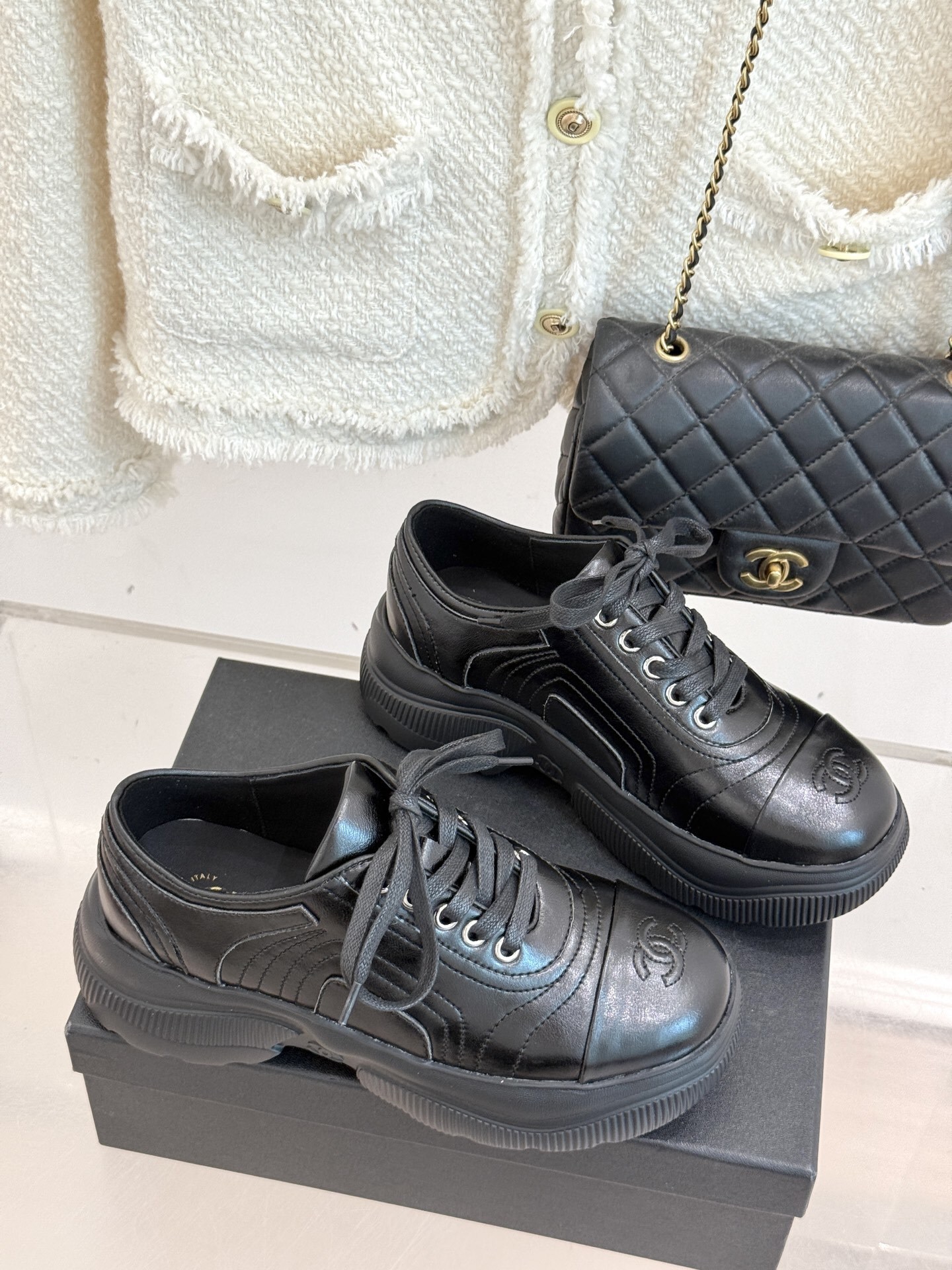 A pair of black Chanel leather platform sneakers with embossed CC logo on a black box, styled with a white tweed jacket and black quilted bag. - Hình 3