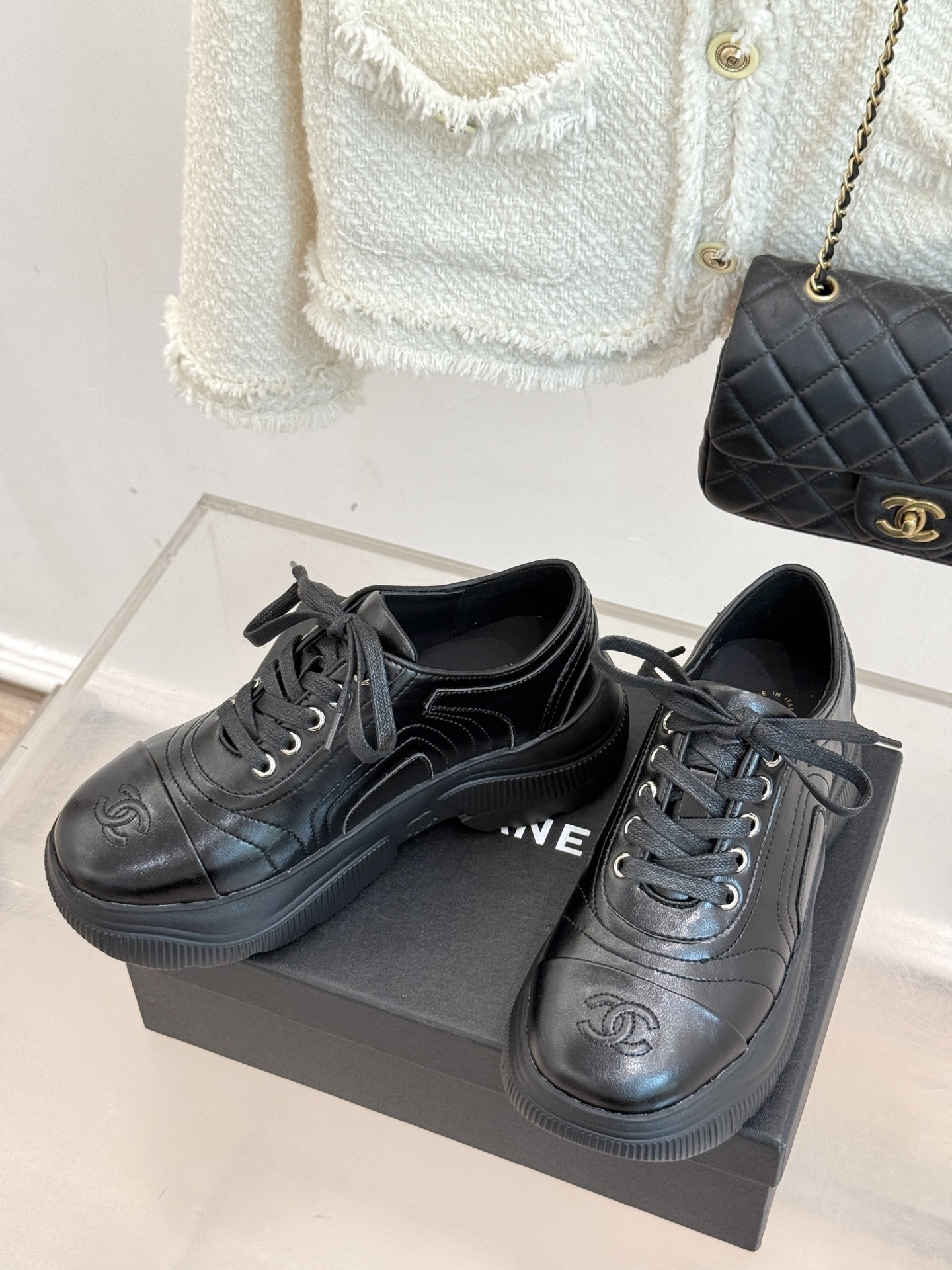 A pair of black Chanel leather platform sneakers with embossed CC logo on a black box, styled with a white tweed jacket and black quilted bag. - Hình 2