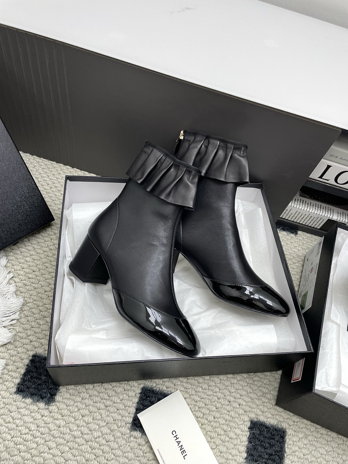 Pair of black Chanel leather ankle boots with ruffle trim and patent leather toe cap on a white surface. - Hình 2