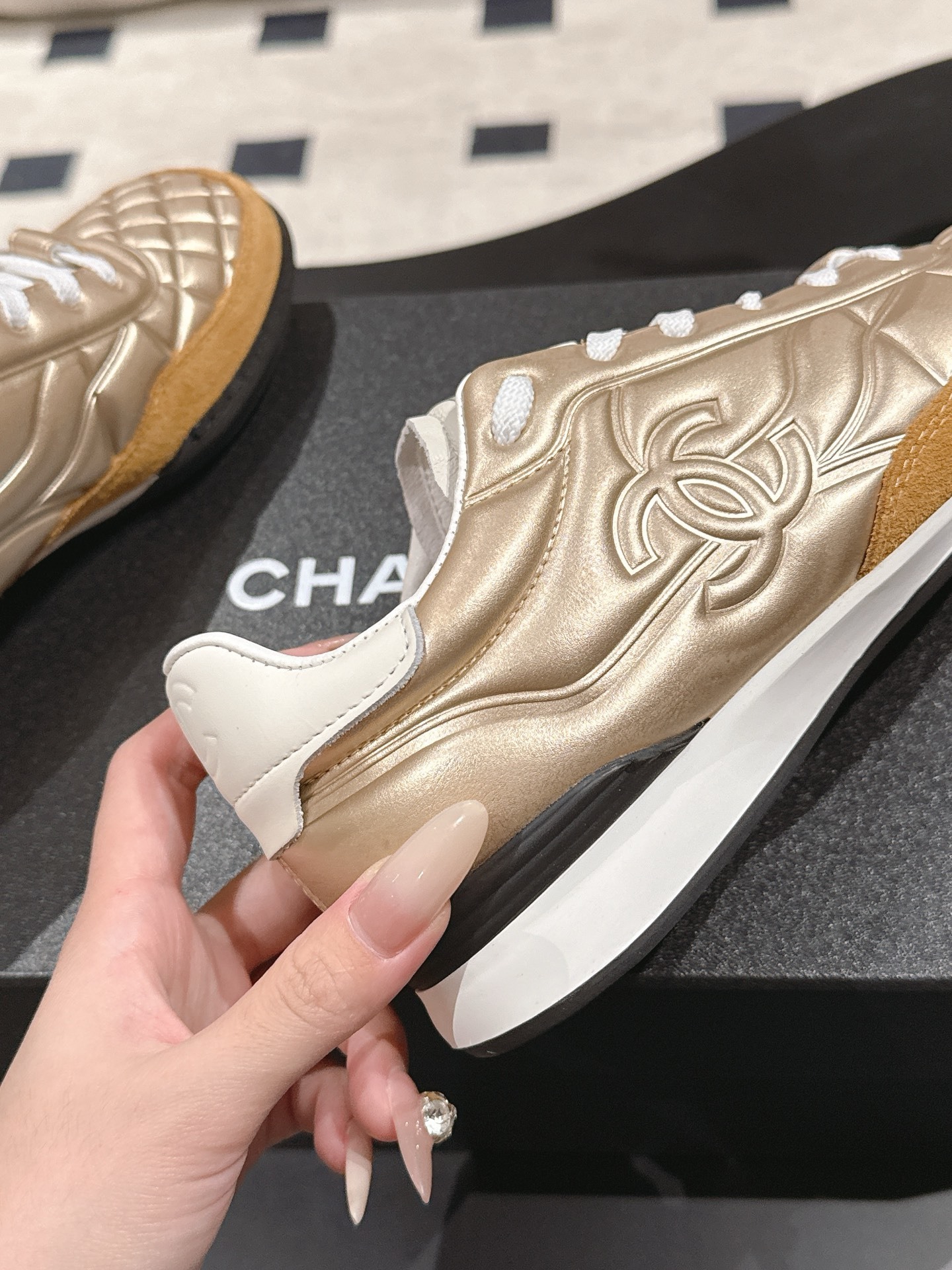 A pair of luxury Chanel metallic gold quilted sneakers with tan suede toe caps resting on a black designer shoe box. - Hình 9