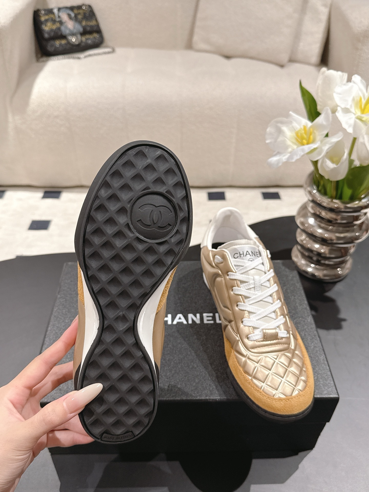 A pair of luxury Chanel metallic gold quilted sneakers with tan suede toe caps resting on a black designer shoe box. - Hình 8