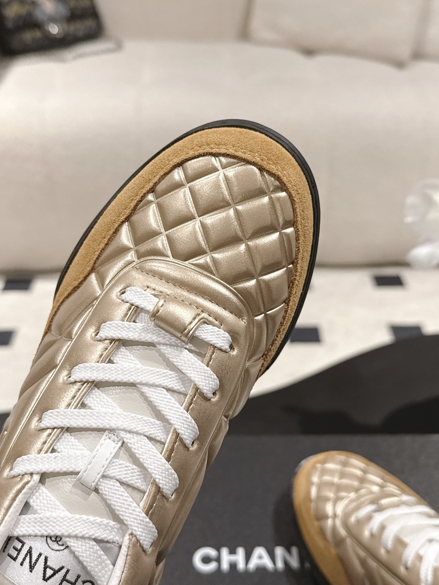 A pair of luxury Chanel metallic gold quilted sneakers with tan suede toe caps resting on a black designer shoe box. - Hình 6
