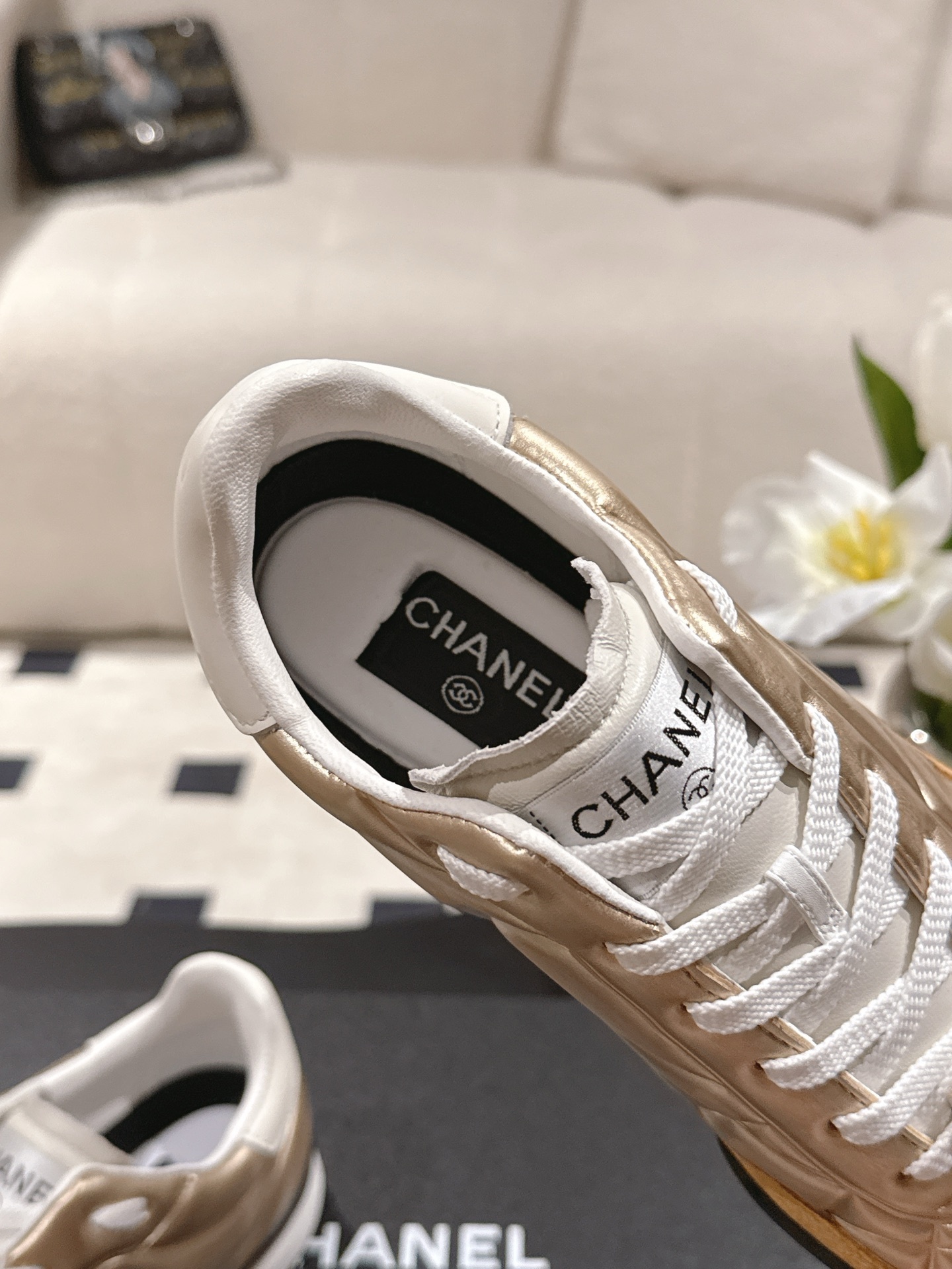 A pair of luxury Chanel metallic gold quilted sneakers with tan suede toe caps resting on a black designer shoe box. - Hình 5