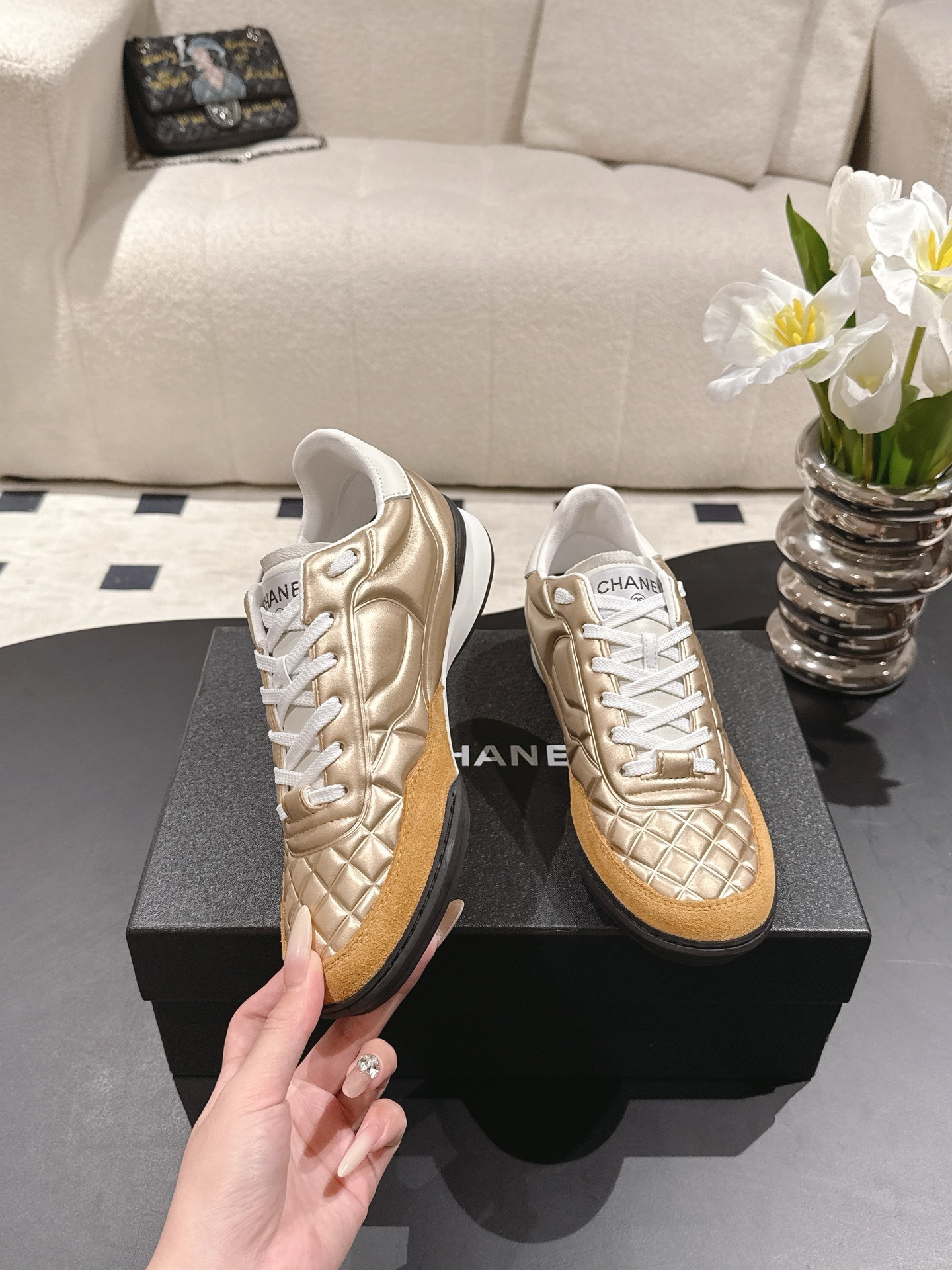 A pair of luxury Chanel metallic gold quilted sneakers with tan suede toe caps resting on a black designer shoe box. - Hình 4