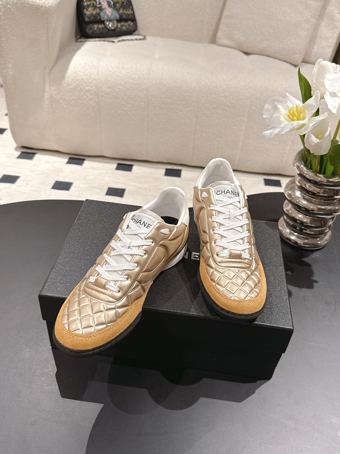 A pair of luxury Chanel metallic gold quilted sneakers with tan suede toe caps resting on a black designer shoe box. - Hình 3