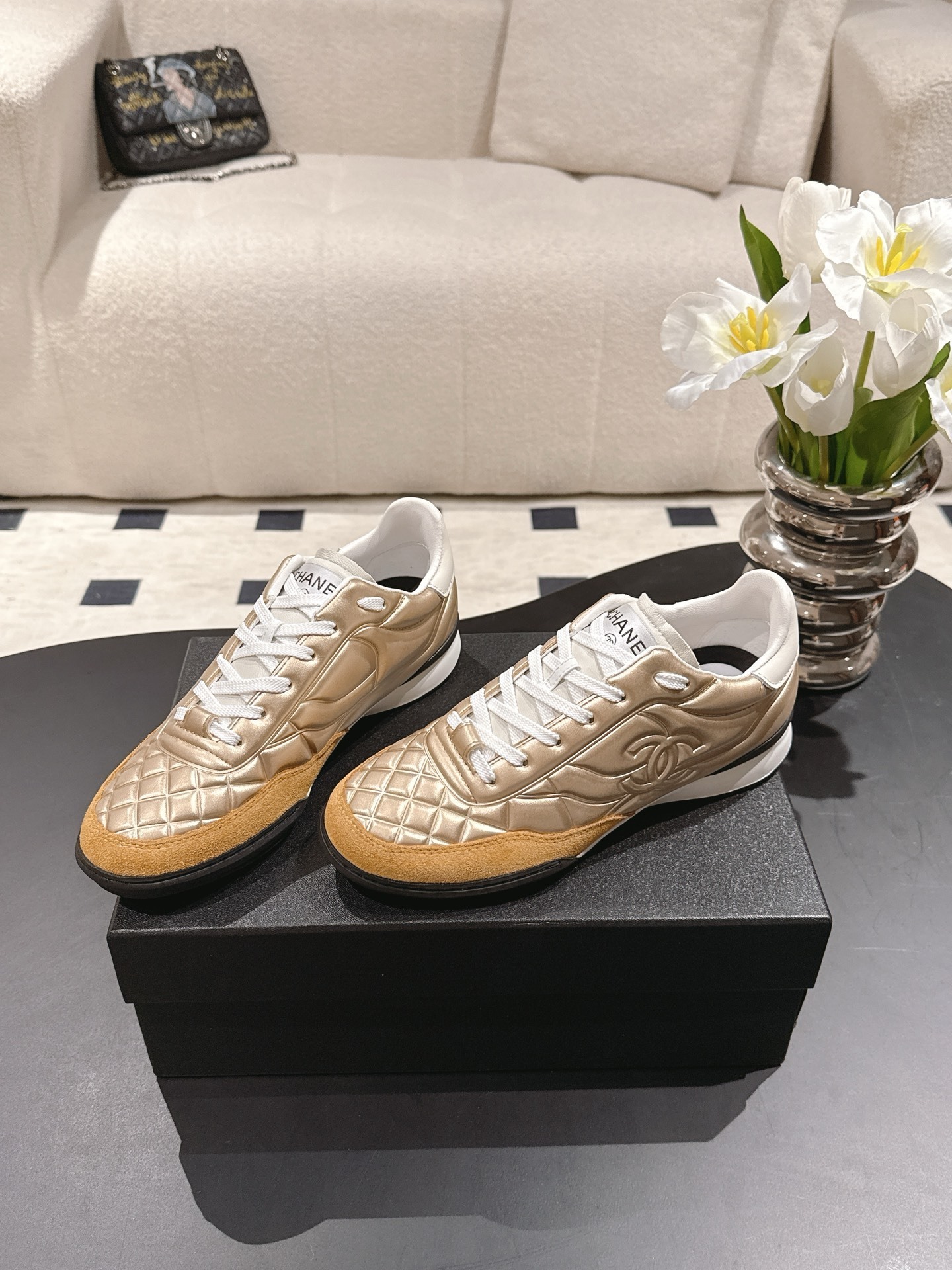 A pair of luxury Chanel metallic gold quilted sneakers with tan suede toe caps resting on a black designer shoe box. - Hình 2