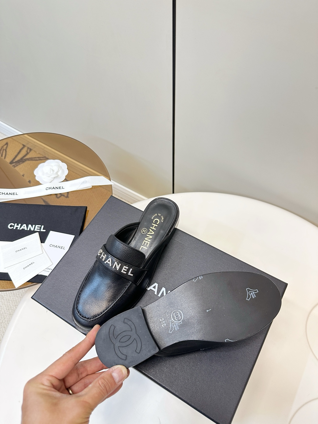 Pair of Chanel black leather backless mules with crystal logo lettering on the strap, resting on a black Chanel box. - Hình 9