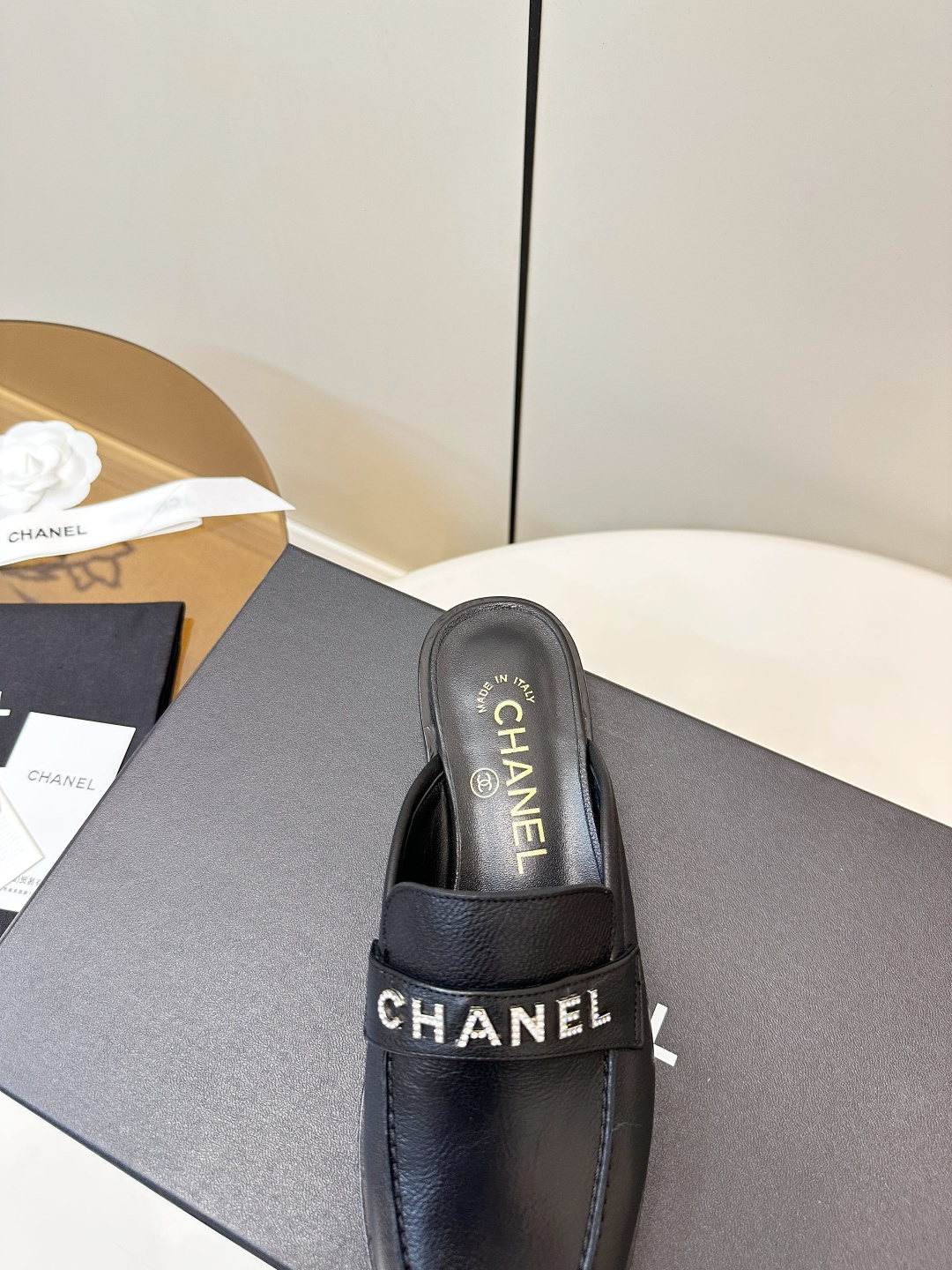 Pair of Chanel black leather backless mules with crystal logo lettering on the strap, resting on a black Chanel box. - Hình 8
