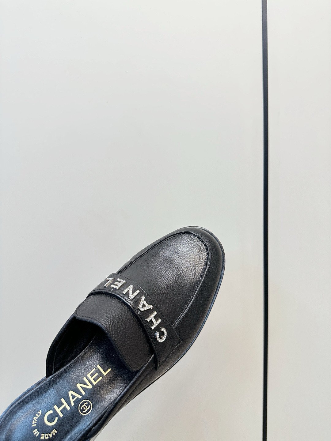 Pair of Chanel black leather backless mules with crystal logo lettering on the strap, resting on a black Chanel box. - Hình 7