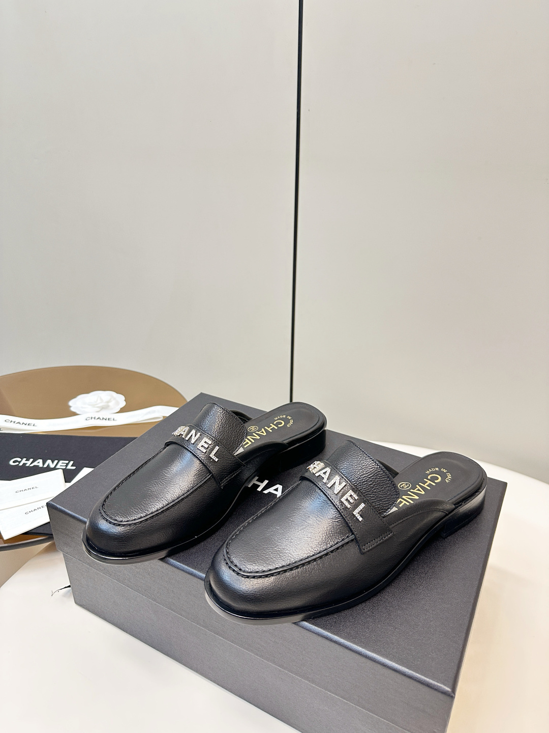 Pair of Chanel black leather backless mules with crystal logo lettering on the strap, resting on a black Chanel box. - Hình 3