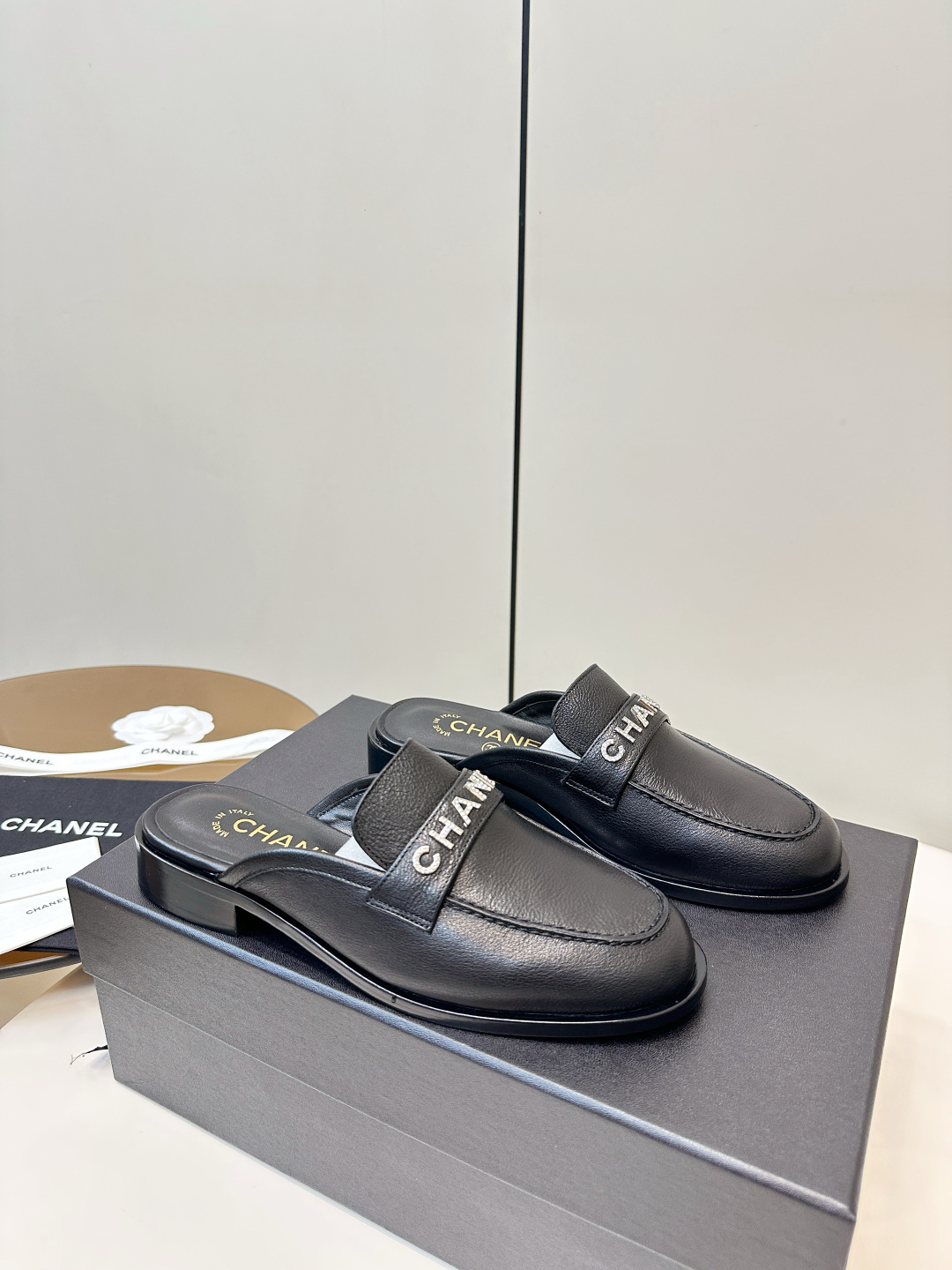 Pair of Chanel black leather backless mules with crystal logo lettering on the strap, resting on a black Chanel box. - Hình 2