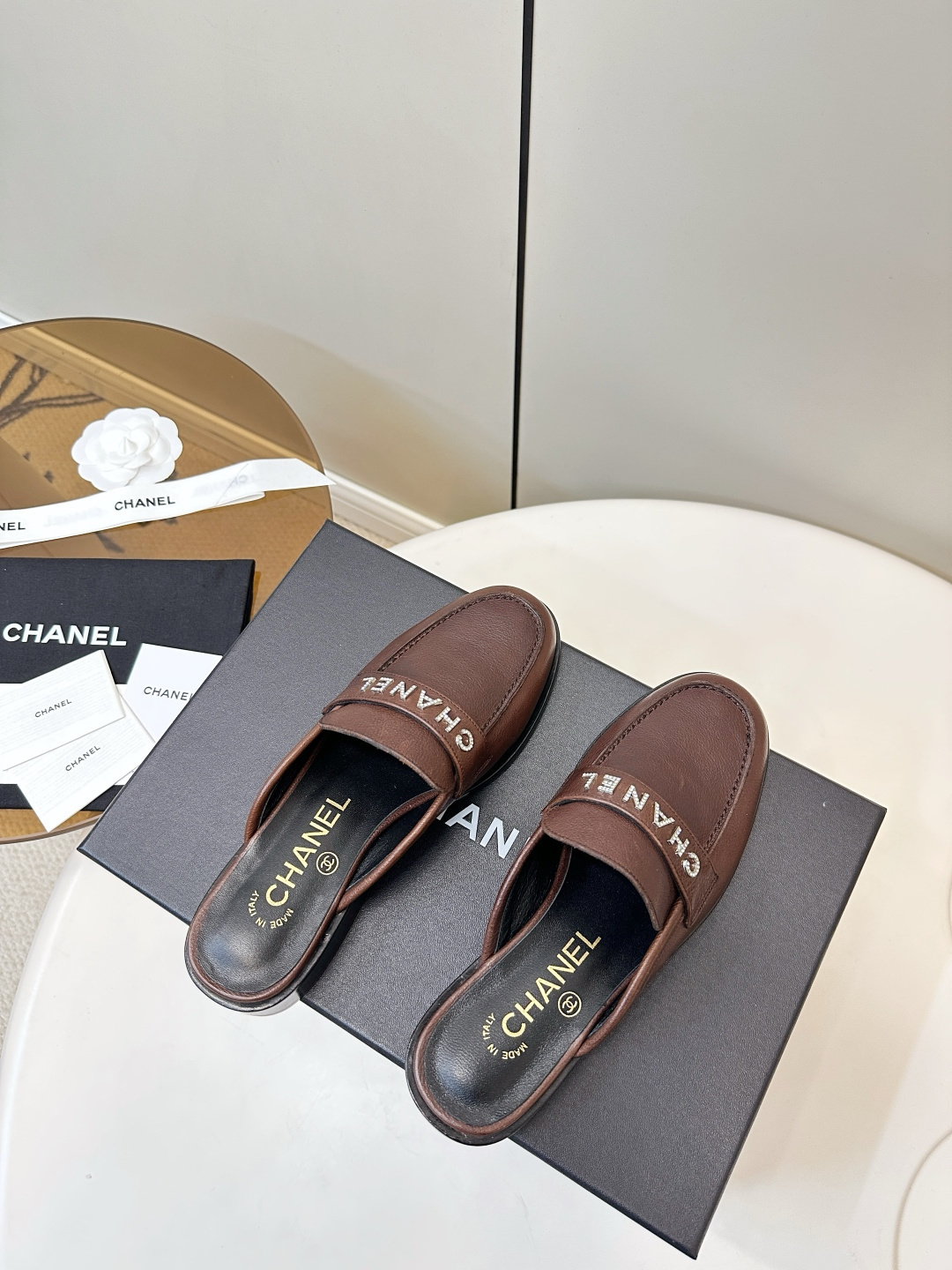 Pair of brown Chanel leather mules with crystal logo lettering displayed on a black box. - Hình 7