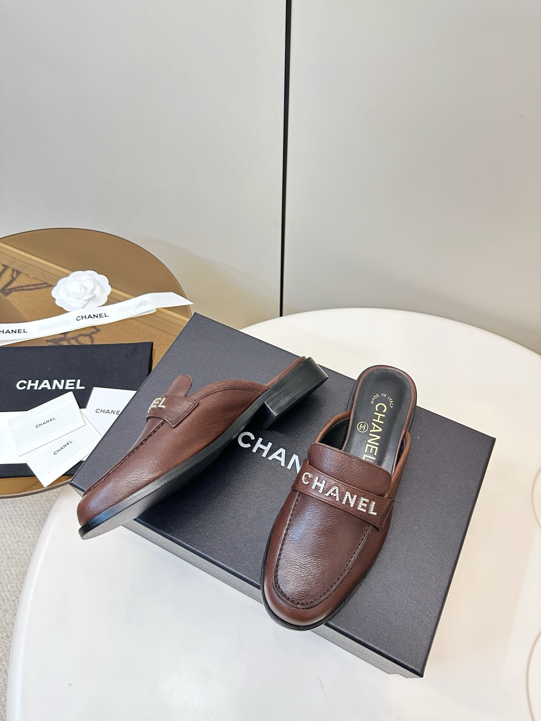 Pair of brown Chanel leather mules with crystal logo lettering displayed on a black box. - Hình 6