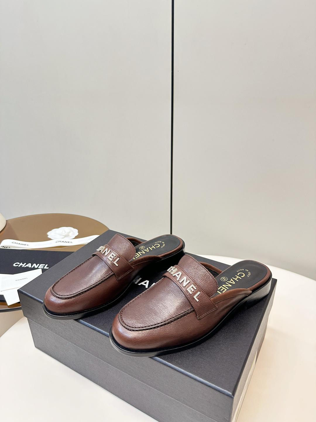 Pair of brown Chanel leather mules with crystal logo lettering displayed on a black box. - Hình 3