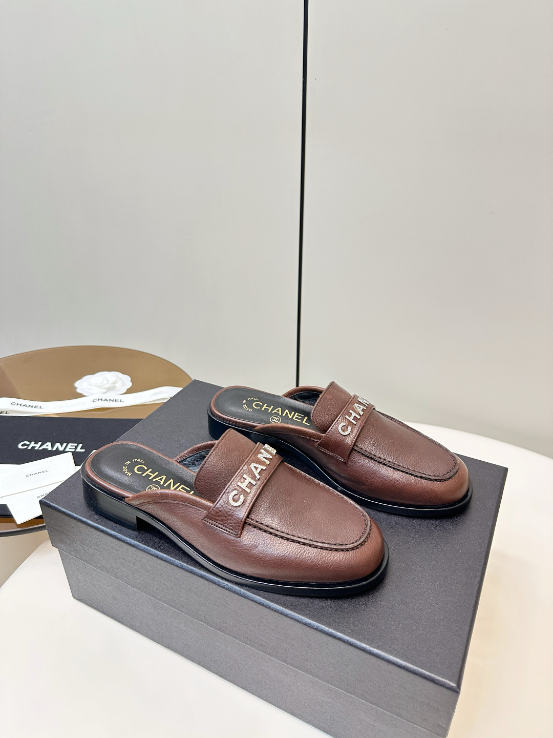 Pair of brown Chanel leather mules with crystal logo lettering displayed on a black box. - Hình 2