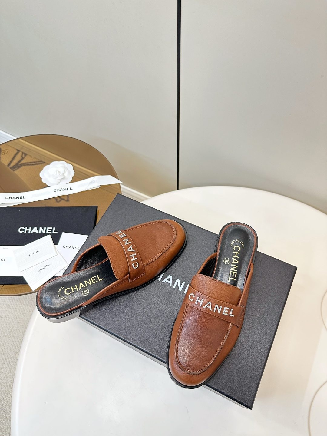 A pair of Chanel brown leather mules with crystal-embellished logo on a black designer box - Hình 8