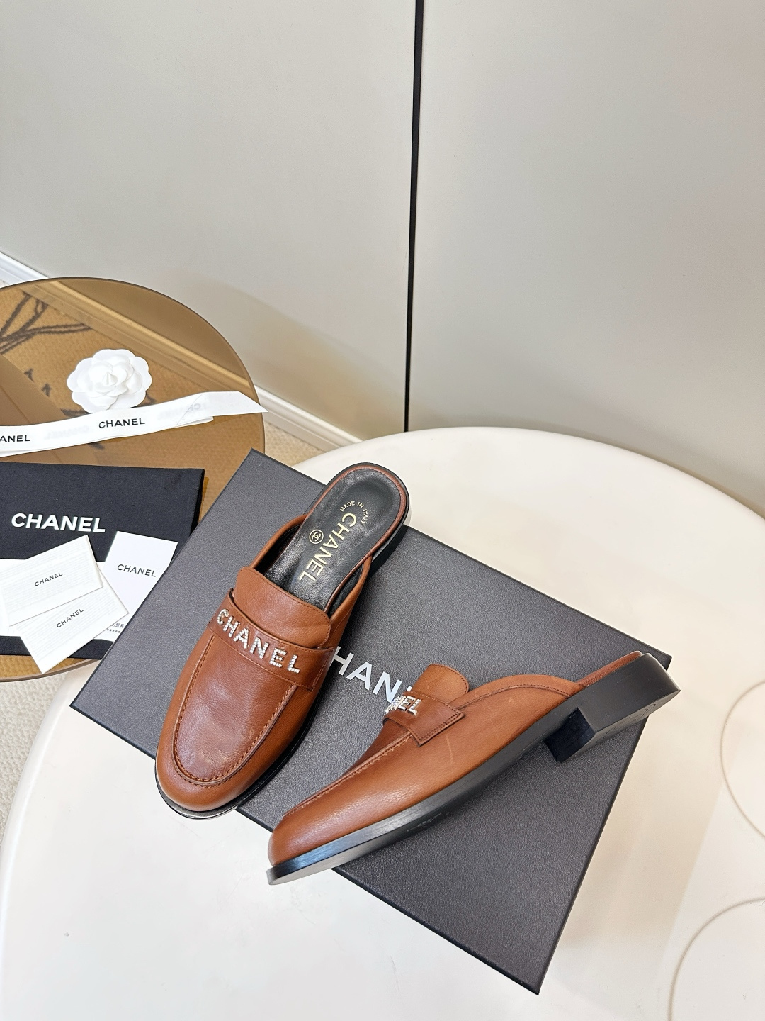 A pair of Chanel brown leather mules with crystal-embellished logo on a black designer box - Hình 7