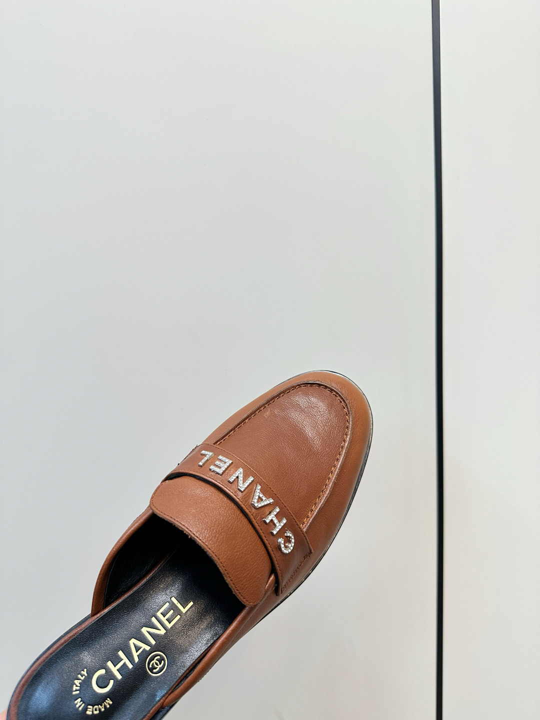 A pair of Chanel brown leather mules with crystal-embellished logo on a black designer box - Hình 6