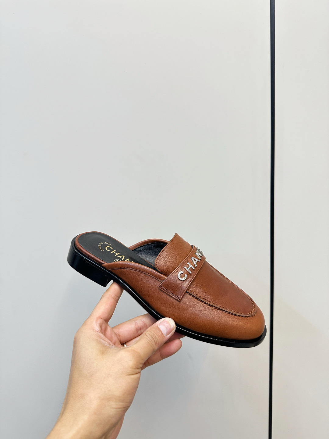 A pair of Chanel brown leather mules with crystal-embellished logo on a black designer box - Hình 5