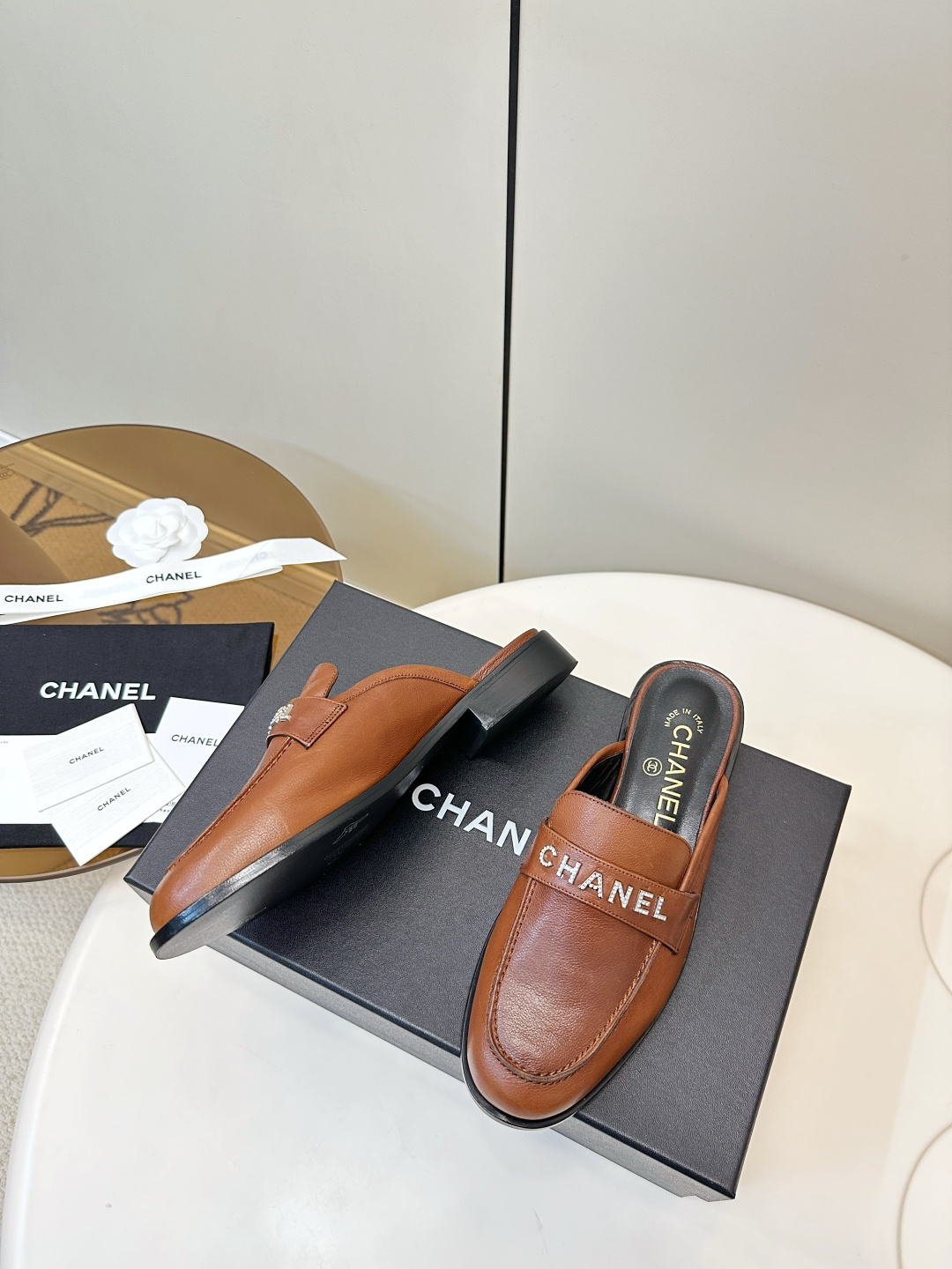 A pair of Chanel brown leather mules with crystal-embellished logo on a black designer box - Hình 4