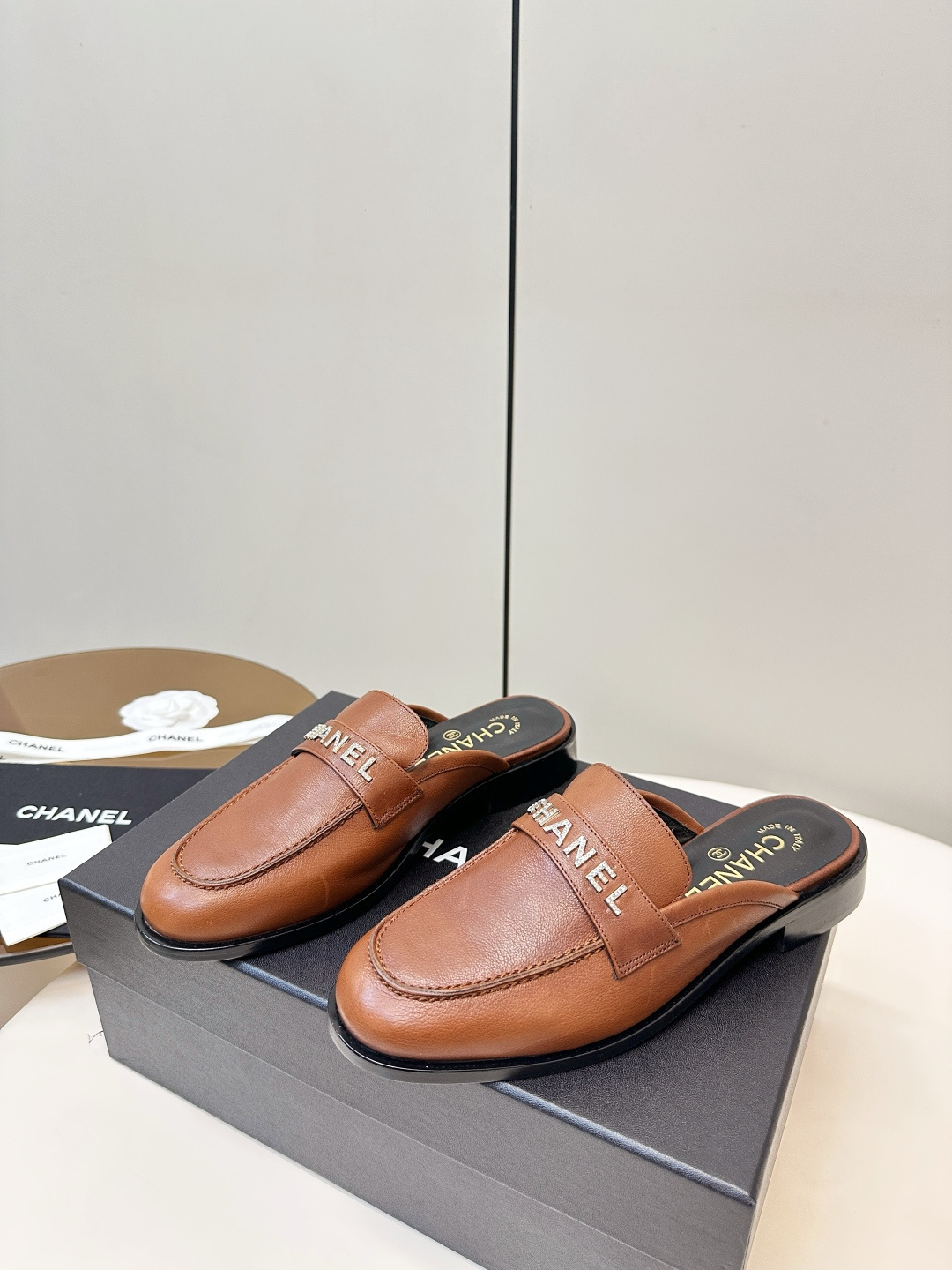 A pair of Chanel brown leather mules with crystal-embellished logo on a black designer box - Hình 3