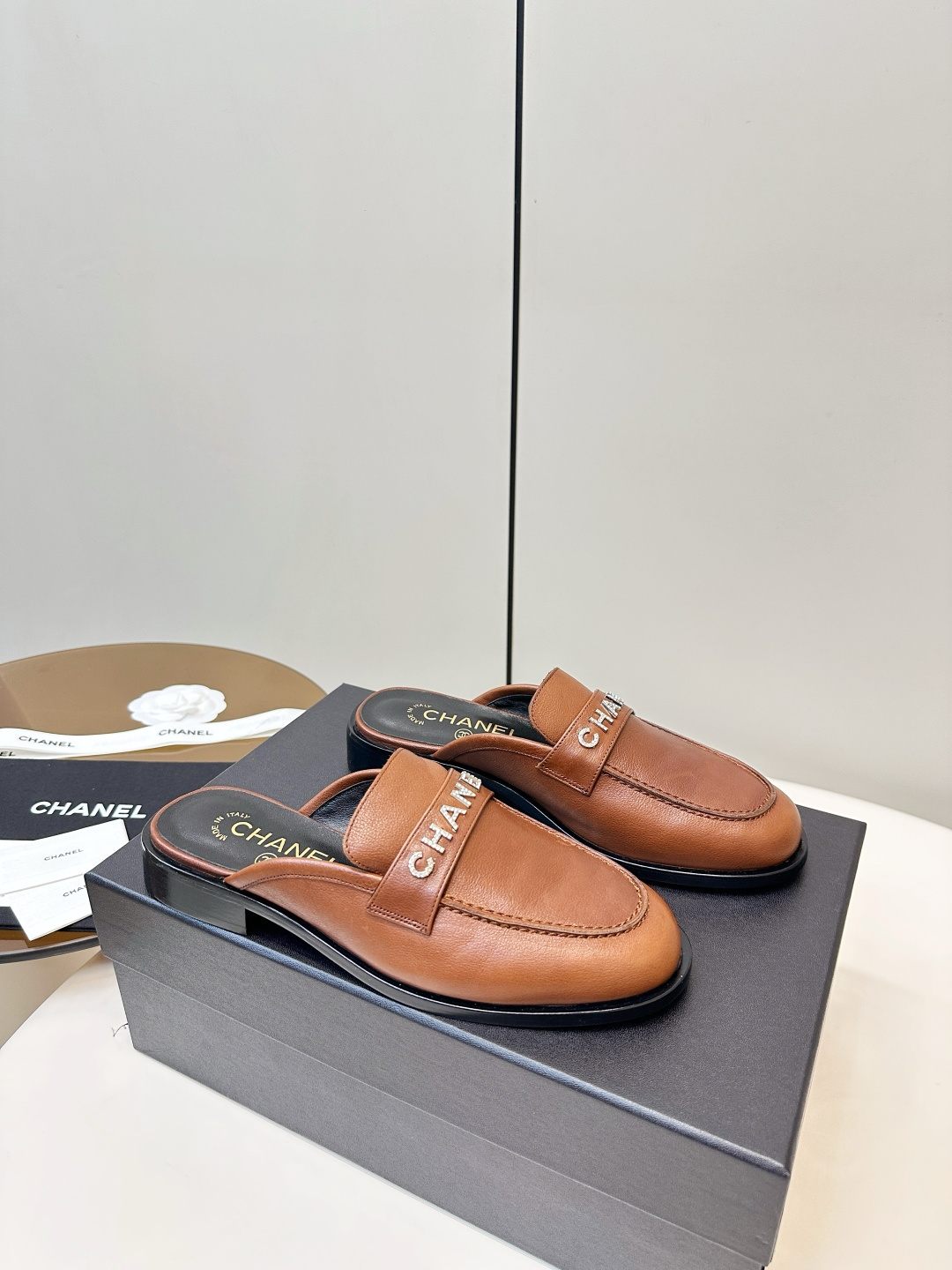 A pair of Chanel brown leather mules with crystal-embellished logo on a black designer box - Hình 2