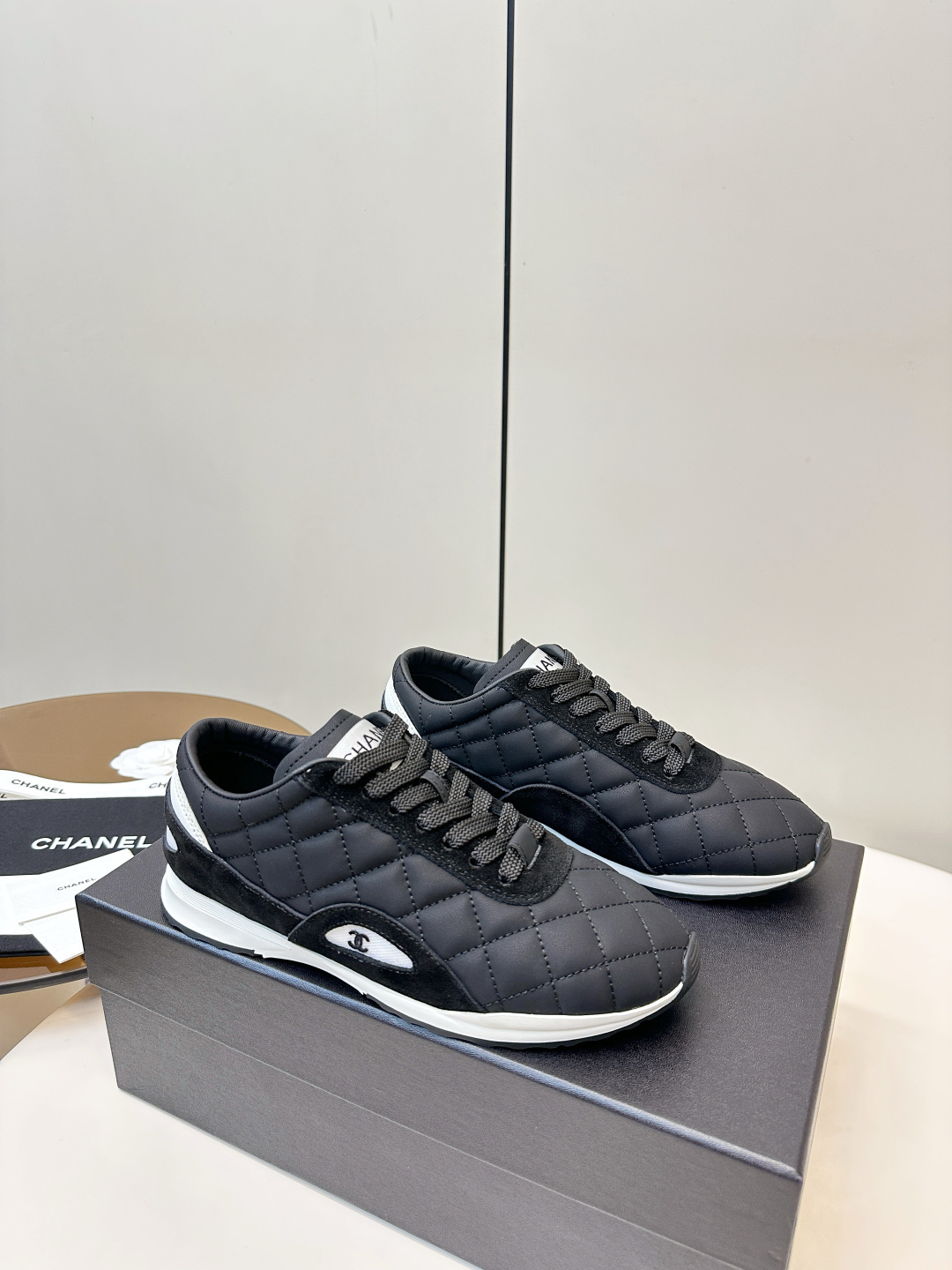 A pair of black Chanel quilted fabric sneakers with white soles and CC logo sitting on a black branded box. - Hình 2