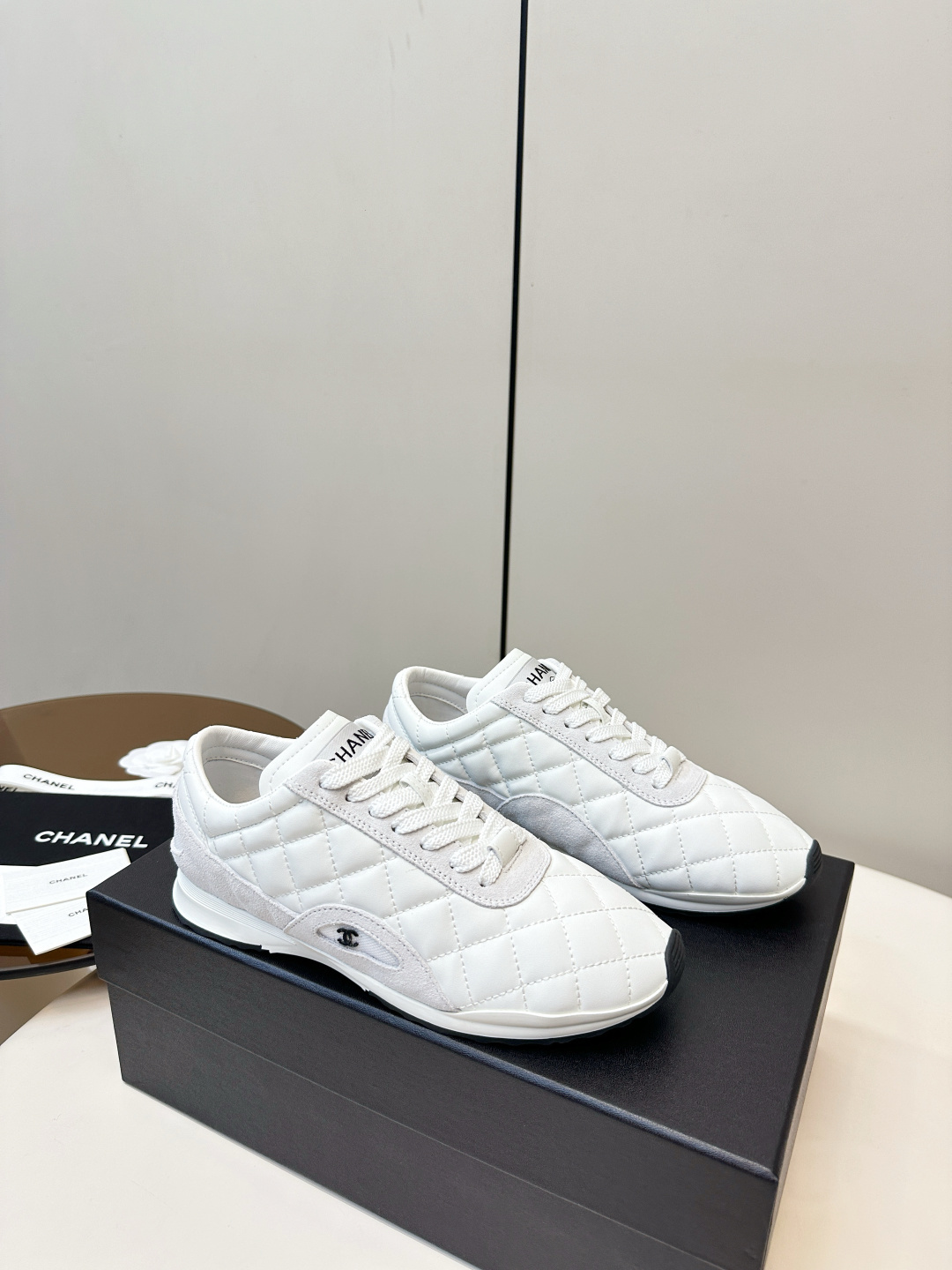 A pair of luxury white Chanel sneakers with a quilted pattern and CC logo resting on a black Chanel shoe box. - Hình 2