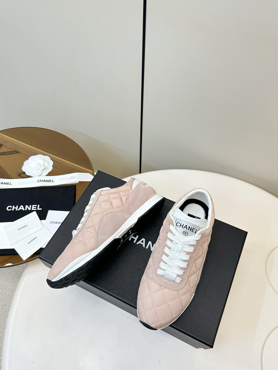 A pair of light pink Chanel quilted sneakers with white laces resting on a black designer box. - Hình 5