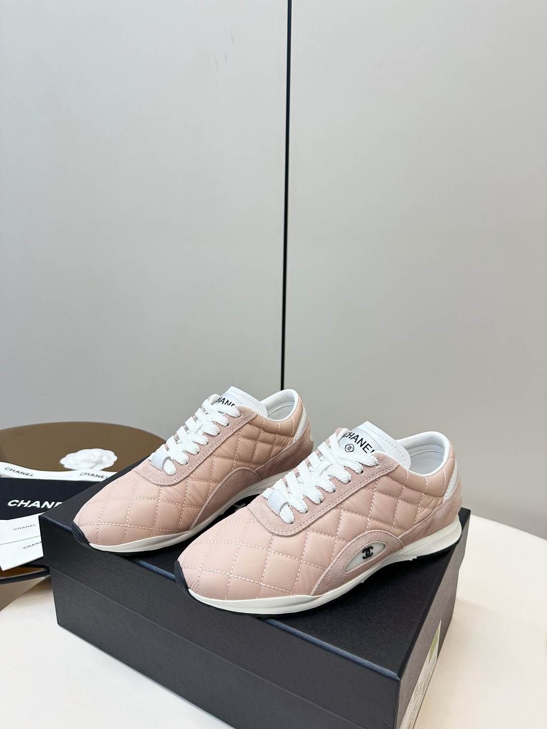 A pair of light pink Chanel quilted sneakers with white laces resting on a black designer box. - Hình 3