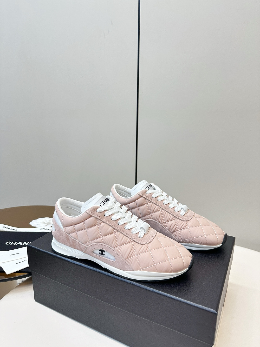 A pair of light pink Chanel quilted sneakers with white laces resting on a black designer box. - Hình 2