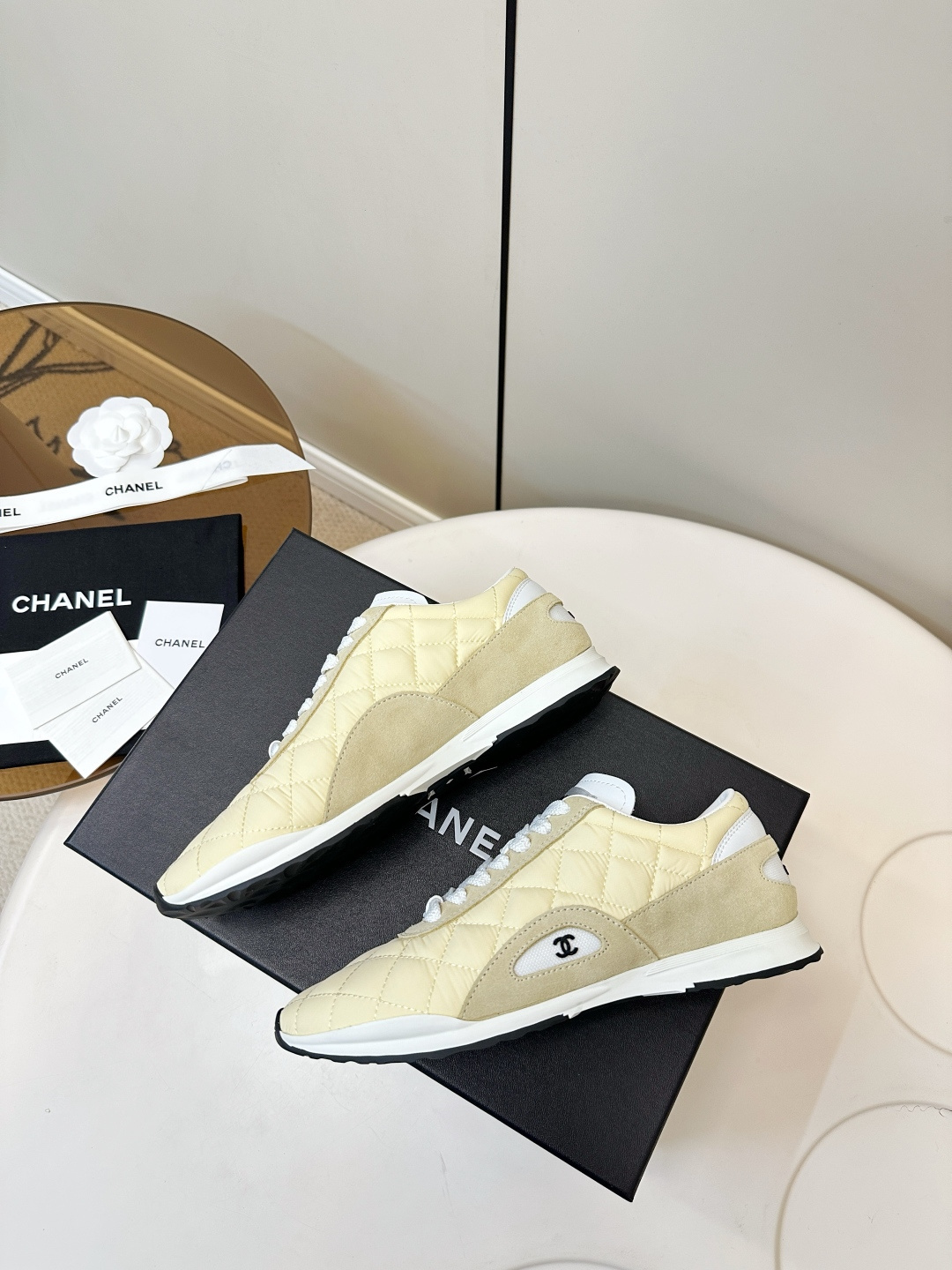 A pair of Chanel pale yellow quilted fabric and suede sneakers resting on a black Chanel designer box with white laces. - Hình 9