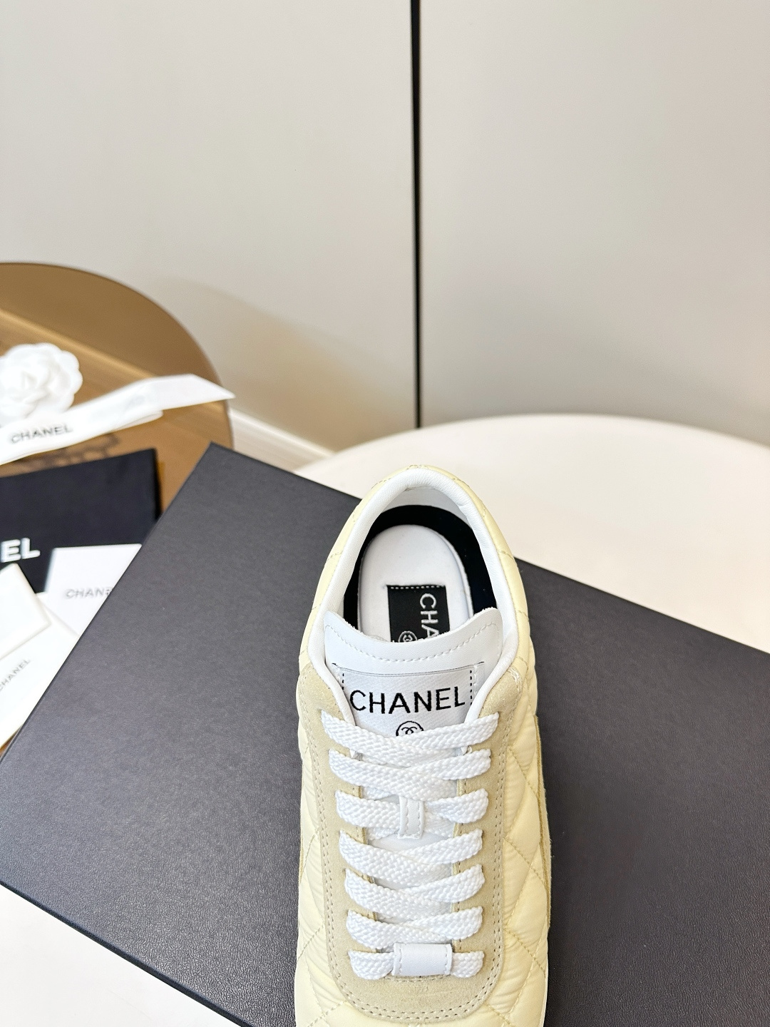 A pair of Chanel pale yellow quilted fabric and suede sneakers resting on a black Chanel designer box with white laces. - Hình 8