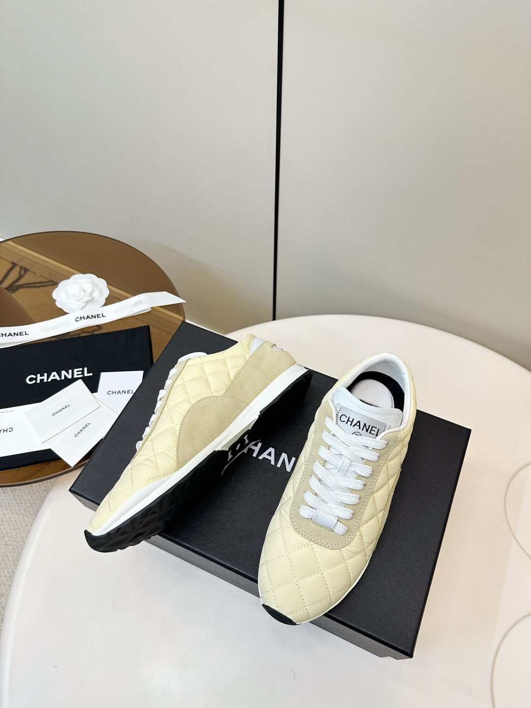 A pair of Chanel pale yellow quilted fabric and suede sneakers resting on a black Chanel designer box with white laces. - Hình 5