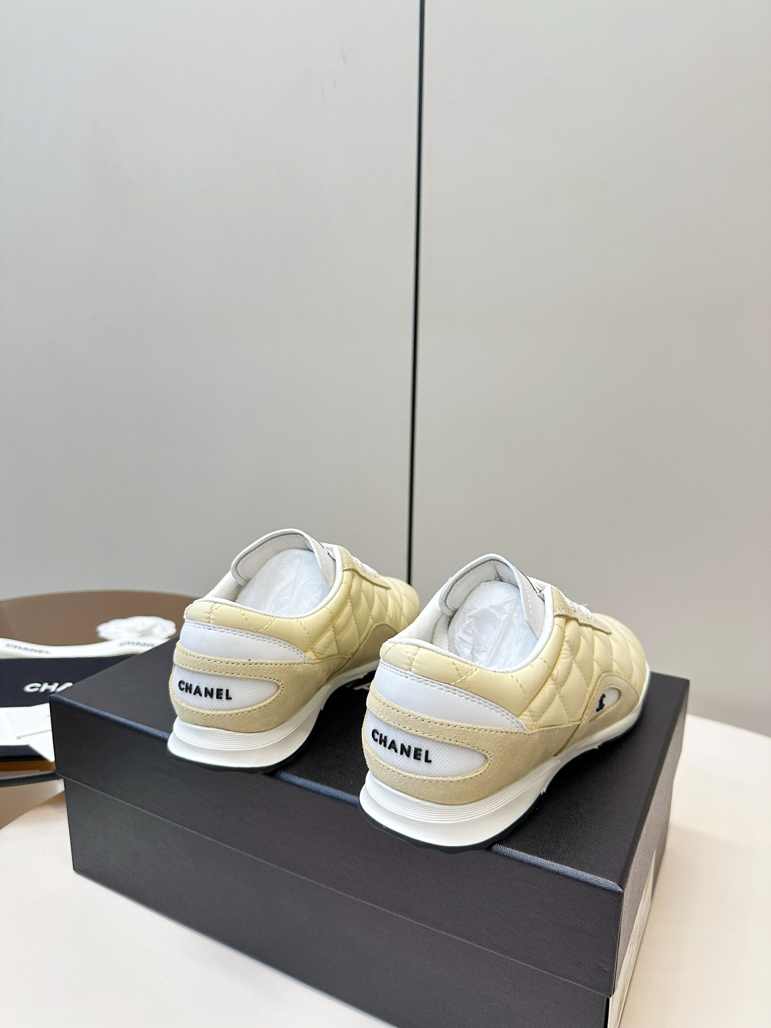 A pair of Chanel pale yellow quilted fabric and suede sneakers resting on a black Chanel designer box with white laces. - Hình 4