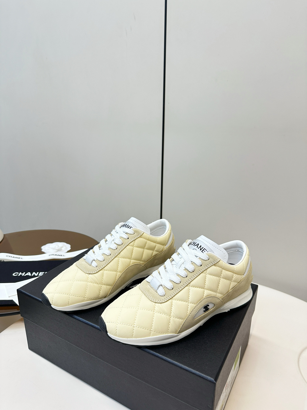 A pair of Chanel pale yellow quilted fabric and suede sneakers resting on a black Chanel designer box with white laces. - Hình 3