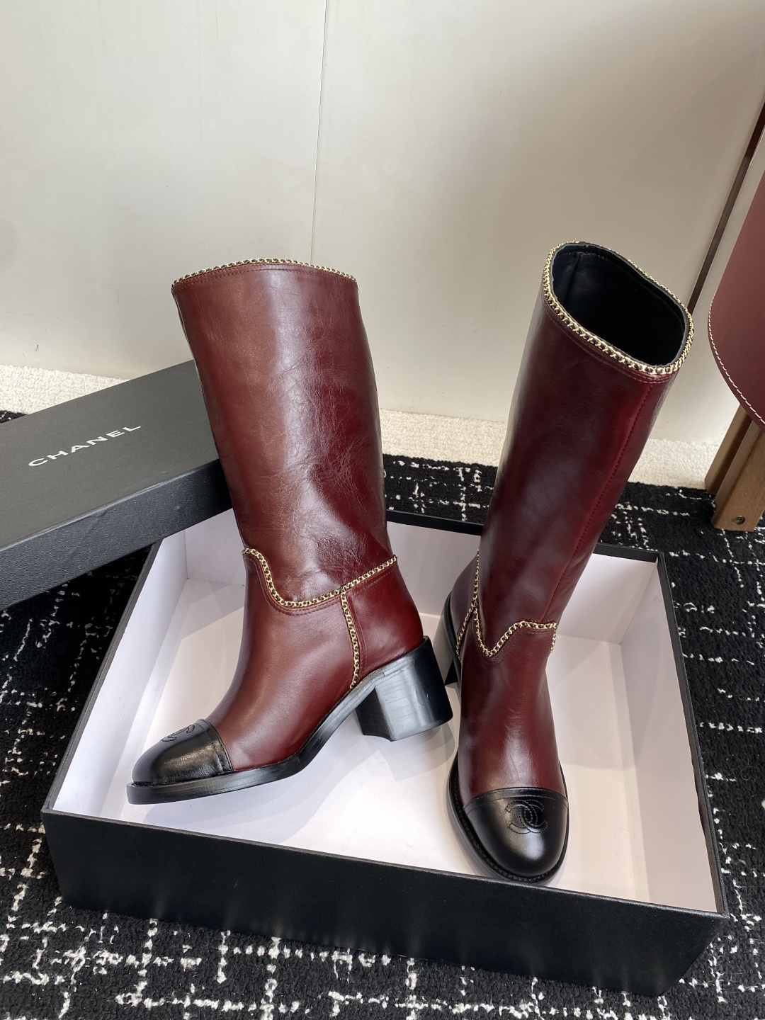 Luxurious Chanel burgundy leather knee-high boots with gold chain trim and black cap-toes displayed on a black box. - Hình 5