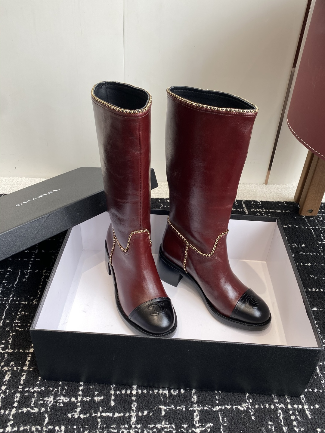 Luxurious Chanel burgundy leather knee-high boots with gold chain trim and black cap-toes displayed on a black box. - Hình 3