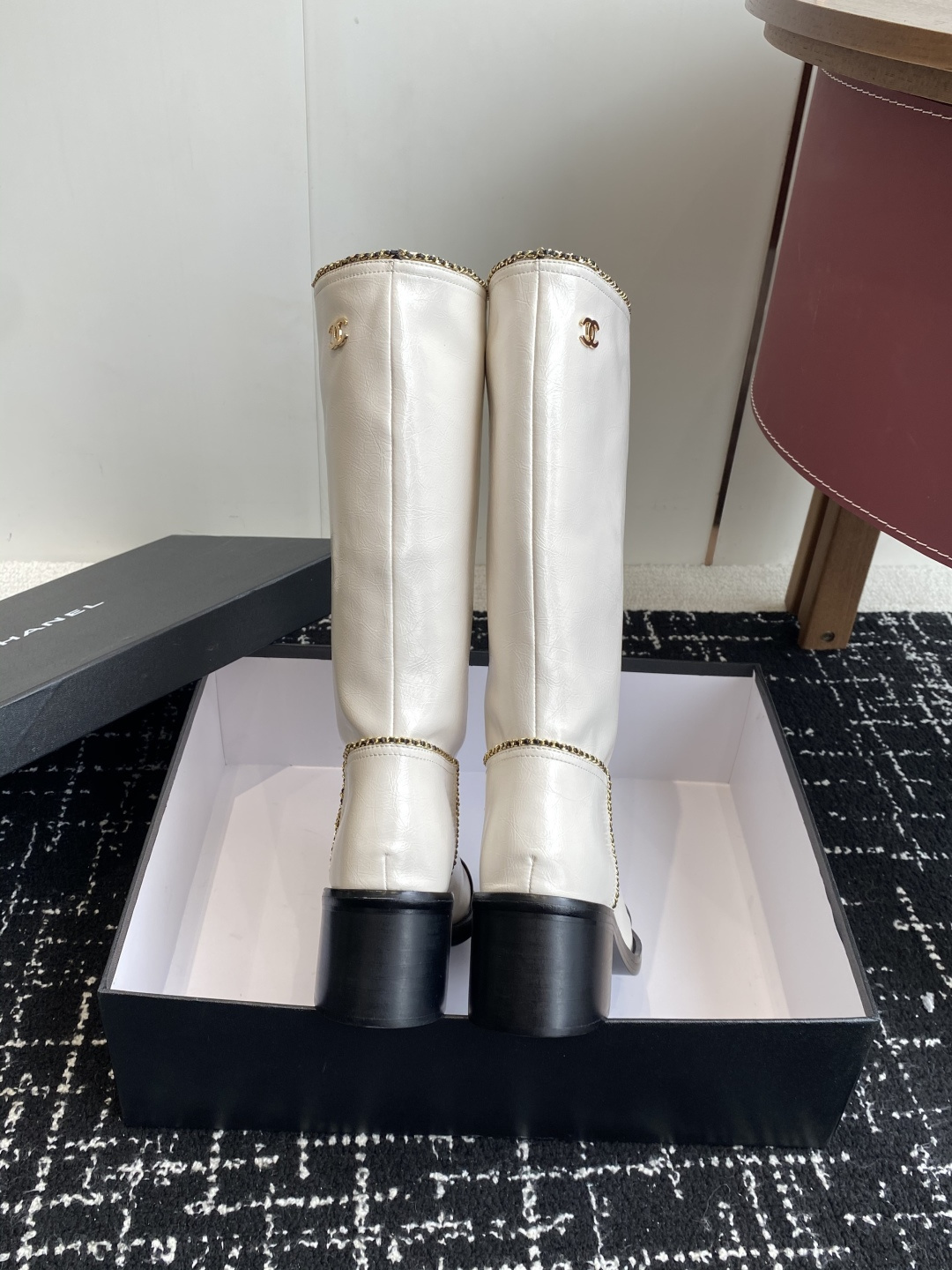 Pair of luxury white and black two-tone knee-high boots with gold chain trim and block heels on a black display box. - Hình 8