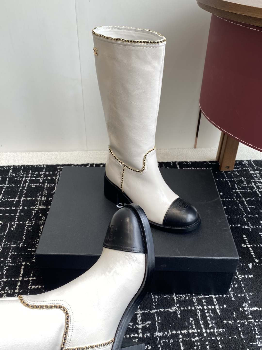 Pair of luxury white and black two-tone knee-high boots with gold chain trim and block heels on a black display box. - Hình 7