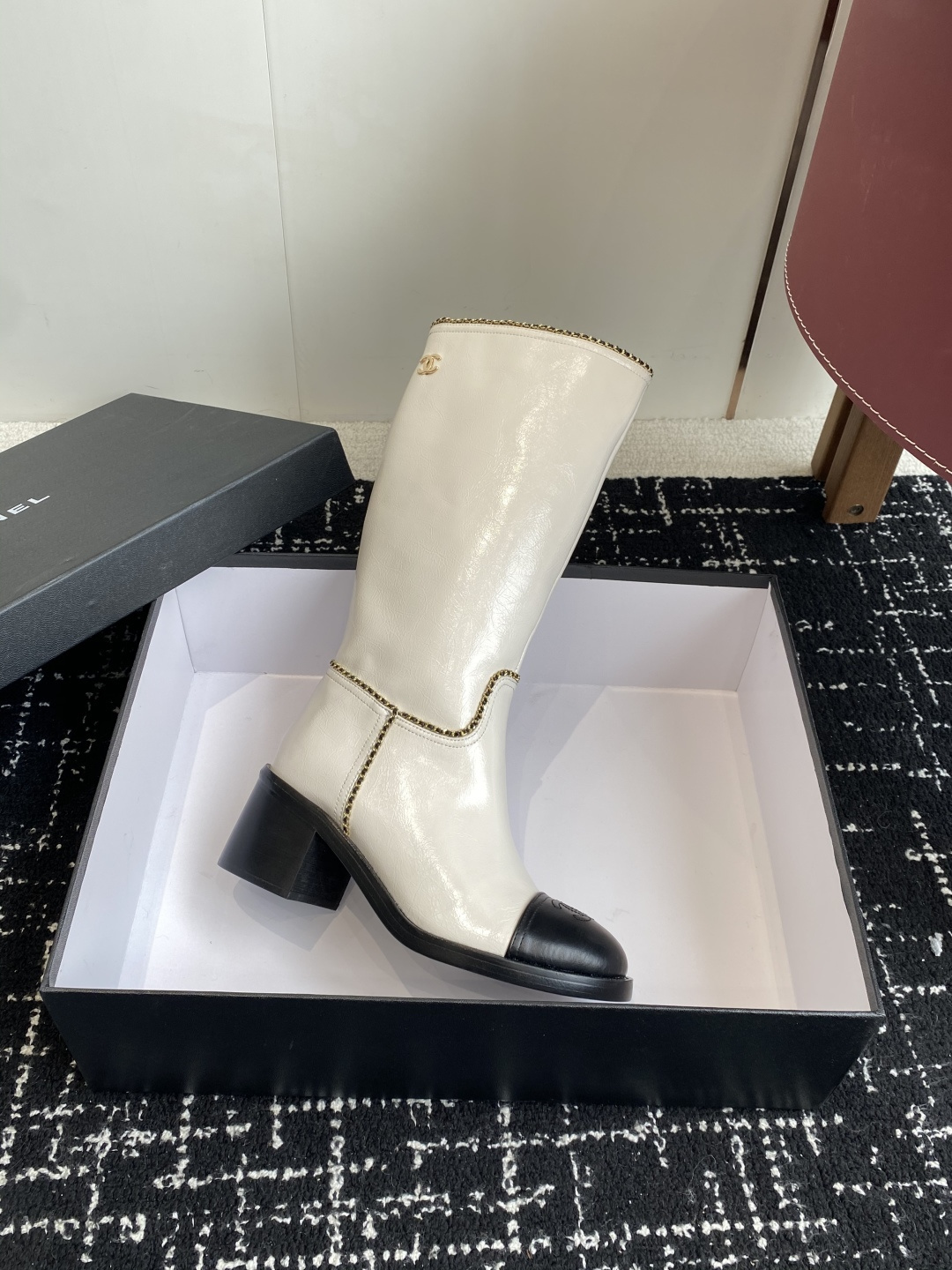Pair of luxury white and black two-tone knee-high boots with gold chain trim and block heels on a black display box. - Hình 6