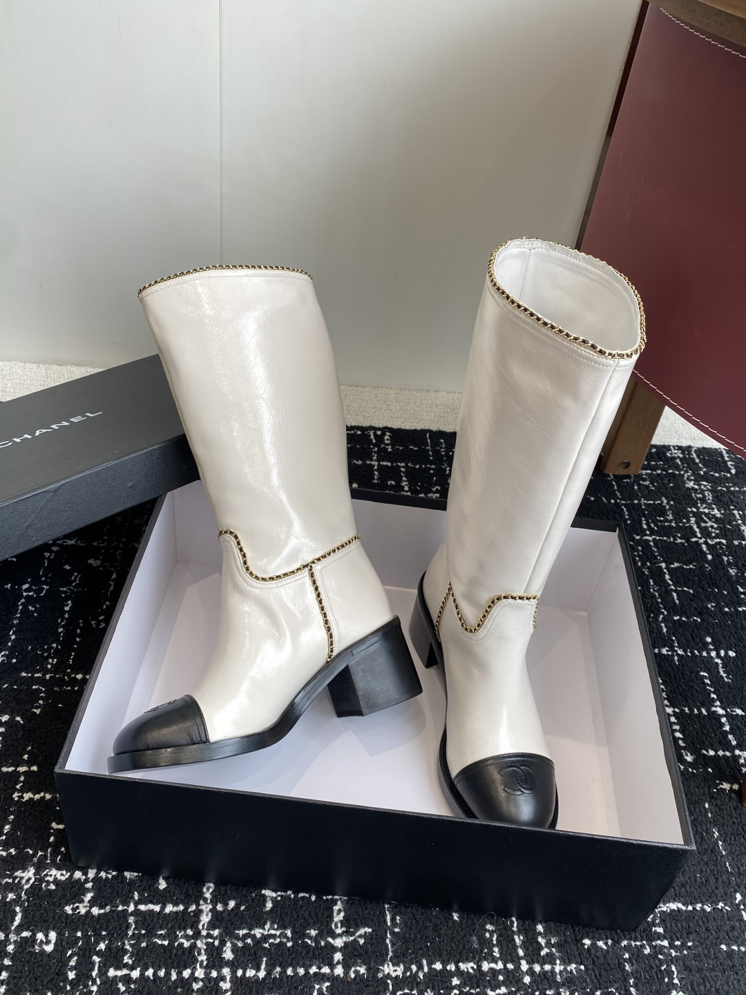 Pair of luxury white and black two-tone knee-high boots with gold chain trim and block heels on a black display box. - Hình 5