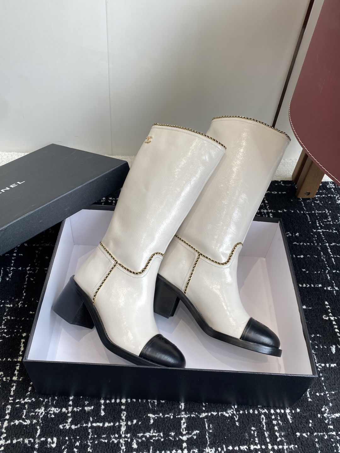 Pair of luxury white and black two-tone knee-high boots with gold chain trim and block heels on a black display box. - Hình 4